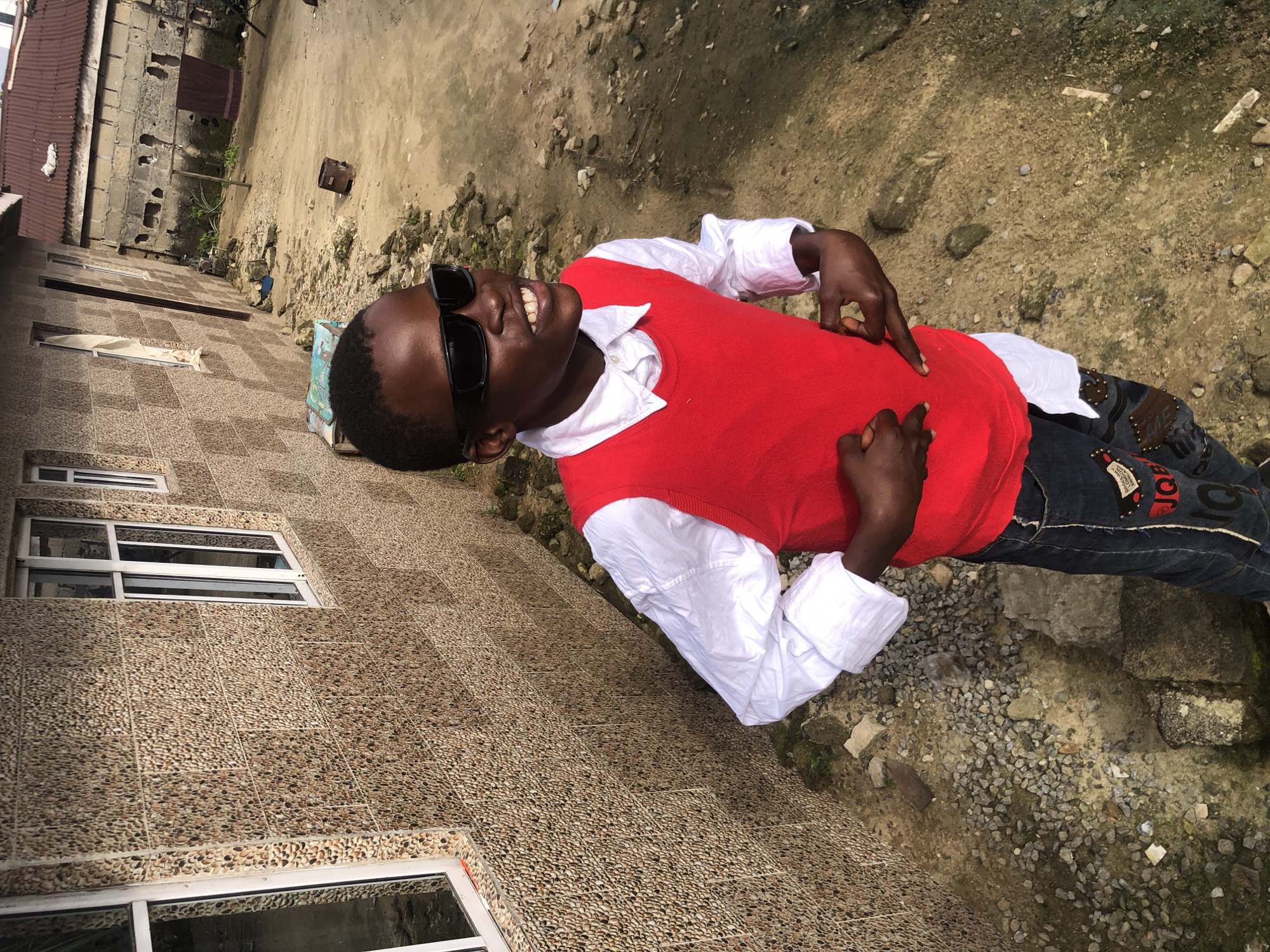 In Ado, Nigeria, a young boy wearing sunglasses, a white collared shirt, a red vest, and jeans stands outside a building. The building's exterior wall is covered in a textured, pebble-like material. In the background, a dirt yard is visible, with scattered debris and a corrugated metal roof on another structure. The boy is looking to his right with a slight smile, and his hands are held in a gesture, with his fingers extended. The overall scene depicts a candid moment in an outdoor urban setting.
