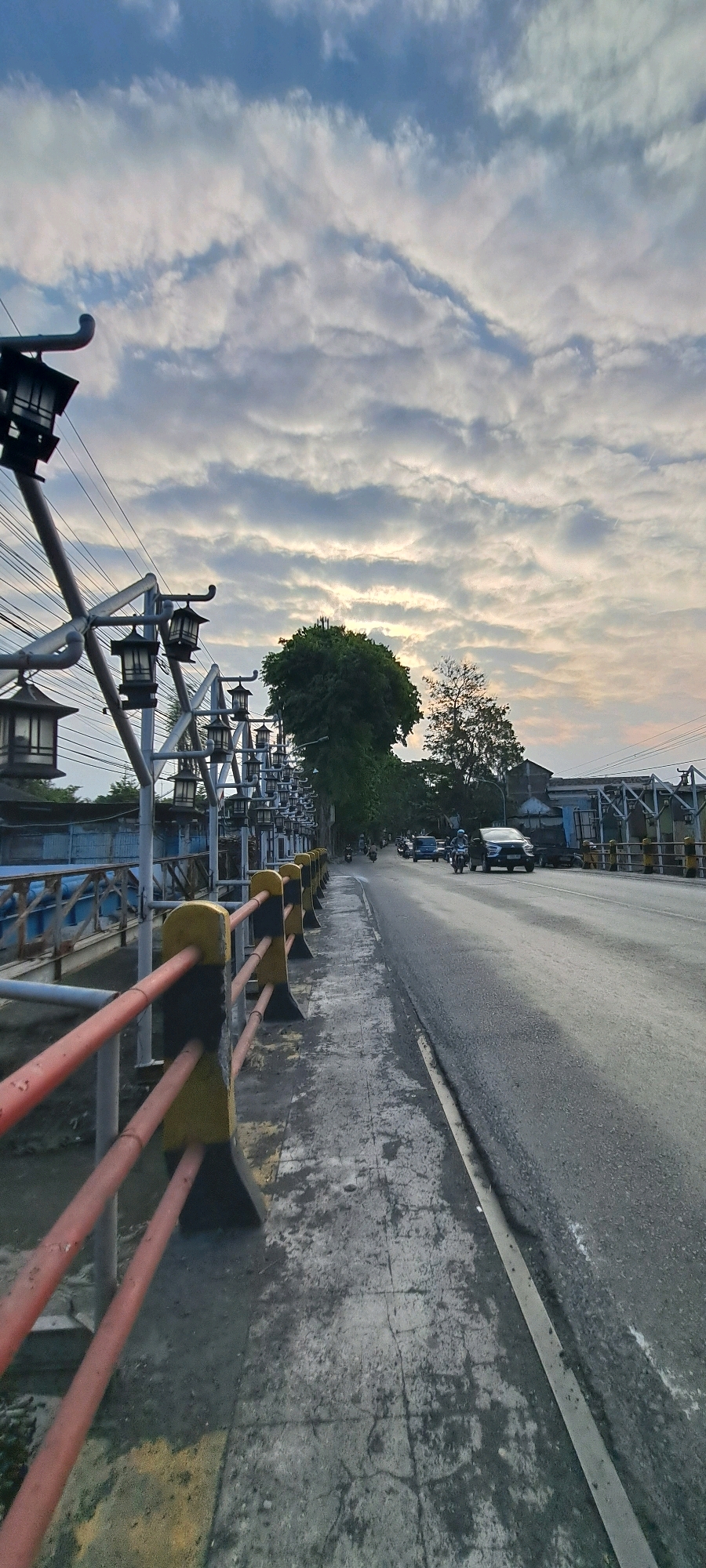 The image depicts a street scene at what appears to be dusk or dawn, possibly on a bridge. The sky is filled with dramatic clouds, hinting at either an impending storm or the aftermath of one, with hints of sunset colors peeking through.
On the left side of the frame, a sidewalk is visible, lined with a railing that features alternating red and yellow painted sections, supported by grey posts. Along the sidewalk, there's a series of ornate street lamps with a dark, angular design, mounted on a metallic structure that stretches into the distance. These lamps are arranged in a repeating pattern, contributing to the aesthetic of the pathway. The sidewalk itself appears weathered, with cracks and discolored patches.
The right side of the image shows a paved road with visible tire marks and some scattered debris. Several vehicles are present on the road, including a dark-colored car and motorcycles, suggesting moderate traffic. The road slopes slightly upwards as it recedes into the distance.
Beyond the sidewalk and railing, a body of water can be glimpsed, likely a river or canal flowing beneath the bridge. Lush green trees, including a prominent large tree, are visible in the background, some of which are climbing over the railing. Buildings and other structures are also present in the distance, indicating an urban environment.
The overall atmosphere is calm, with the streetlights yet to be illuminated, and the natural light from the sky dominating the scene. The image appears to be taken from a low angle, looking down the length of the bridge.
Based on the presence of the distinctive street lamps and the architectural style, this scene is identifiable as the bridge over the river in the city of Pasuruan, Indonesia. The lamps themselves are a characteristic feature of this location, often referred to as "Gerbang Kota" or the city gate bridge.