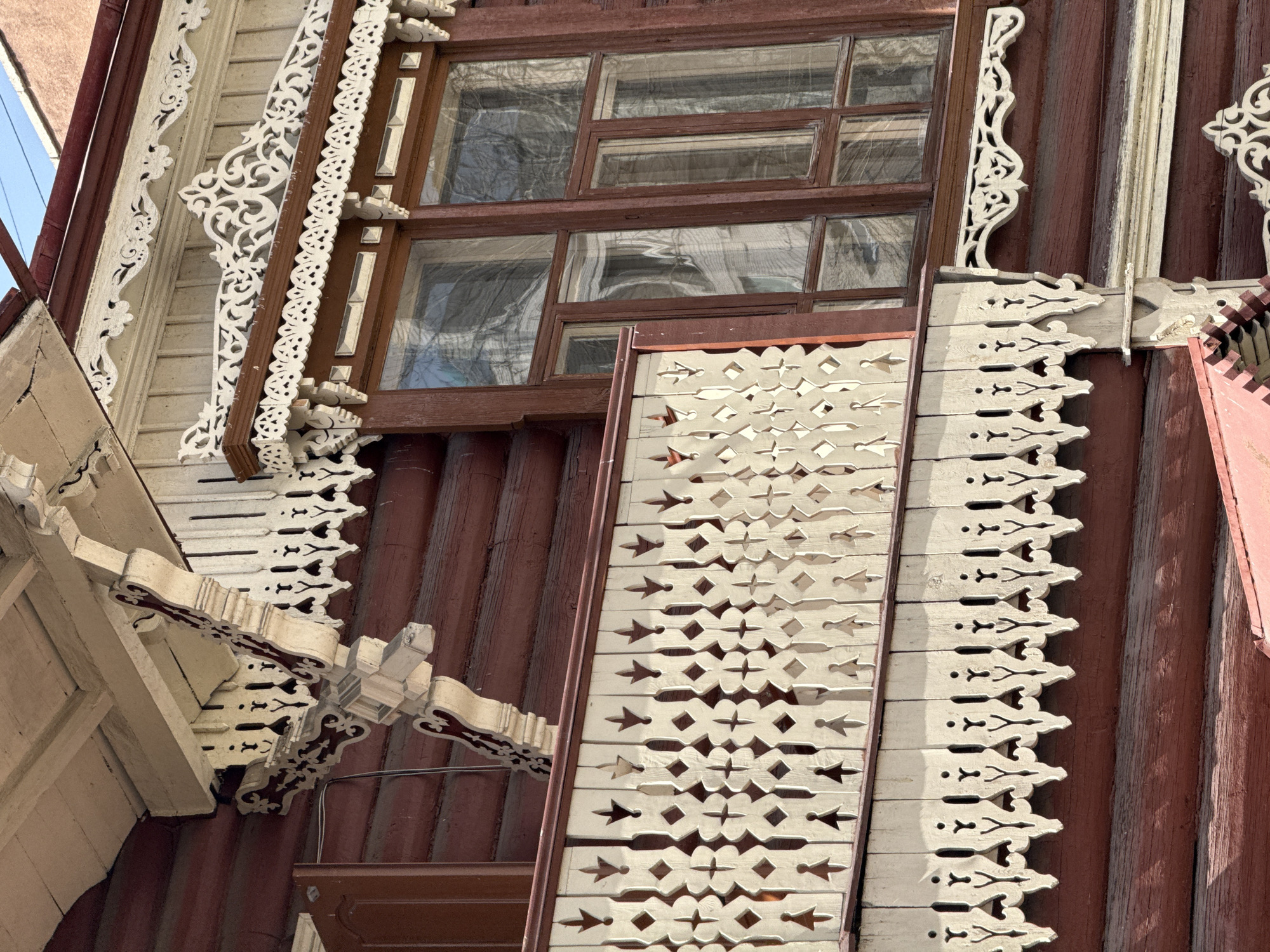 The image captures a close-up view of a section of an elaborately decorated wooden building in Chita, Russia. The building's exterior features rich, dark reddish-brown vertical wooden planks. An old, multi-paned window with a dark brown frame is visible, reflecting the environment outside. Decorative white wooden trim adorns the building around the window and eaves. The trim exhibits intricate, repetitive patterns cut into the wood, creating an openwork effect. There is an angular, cream-colored, decorated eave detail at the bottom-left of the frame. The lighting suggests a sunny day.