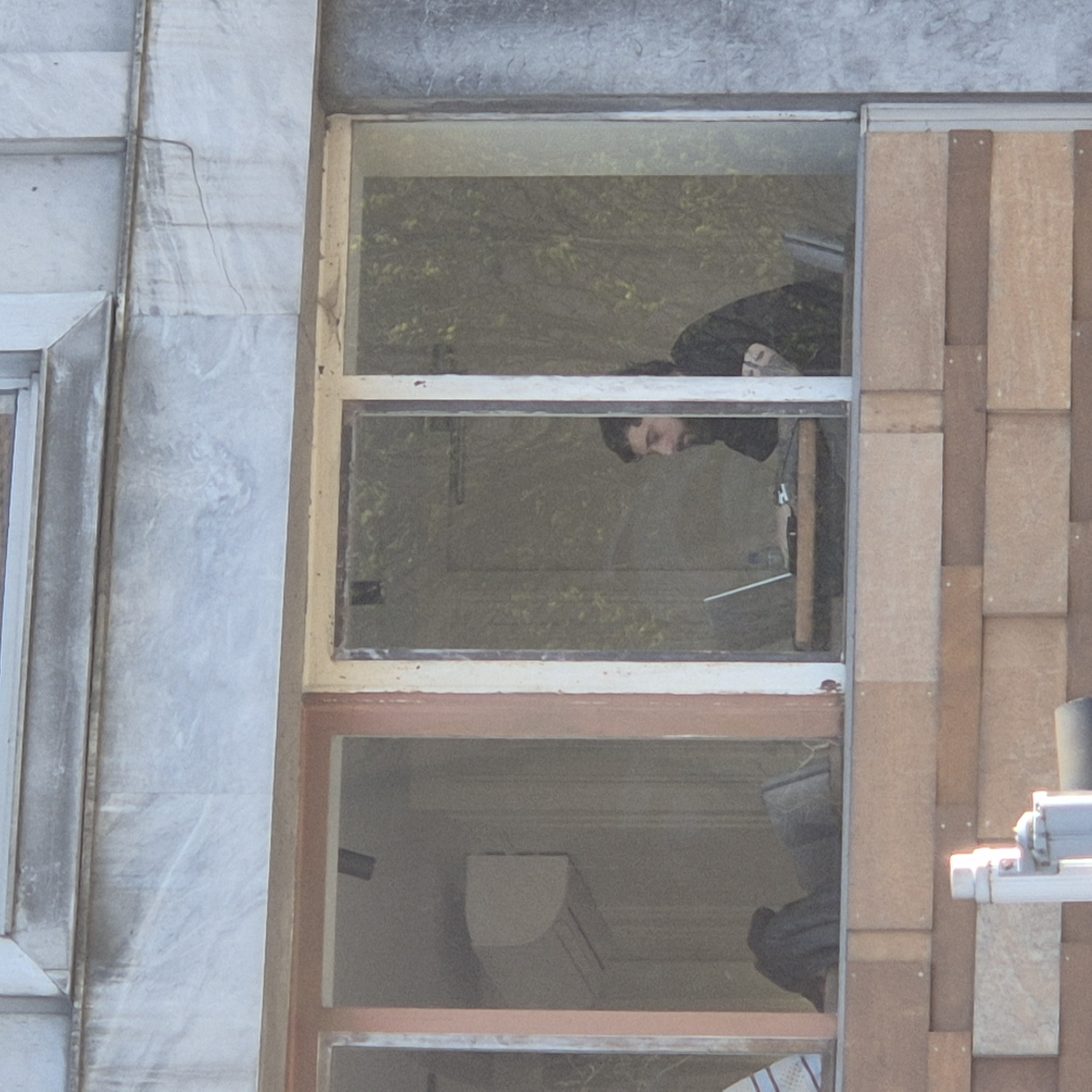 The image shows a man with dark hair and a beard looking out of an open window in a building. The window is part of a larger window unit with three panes. The building's exterior is constructed with a combination of materials; it has a grey stone finish to the left of the window and light brown rectangular tiles to the right. There is an air conditioning unit visible in the bottom window pane. Outside the window, another building and some trees are visible. The location is Strasbourg, France.