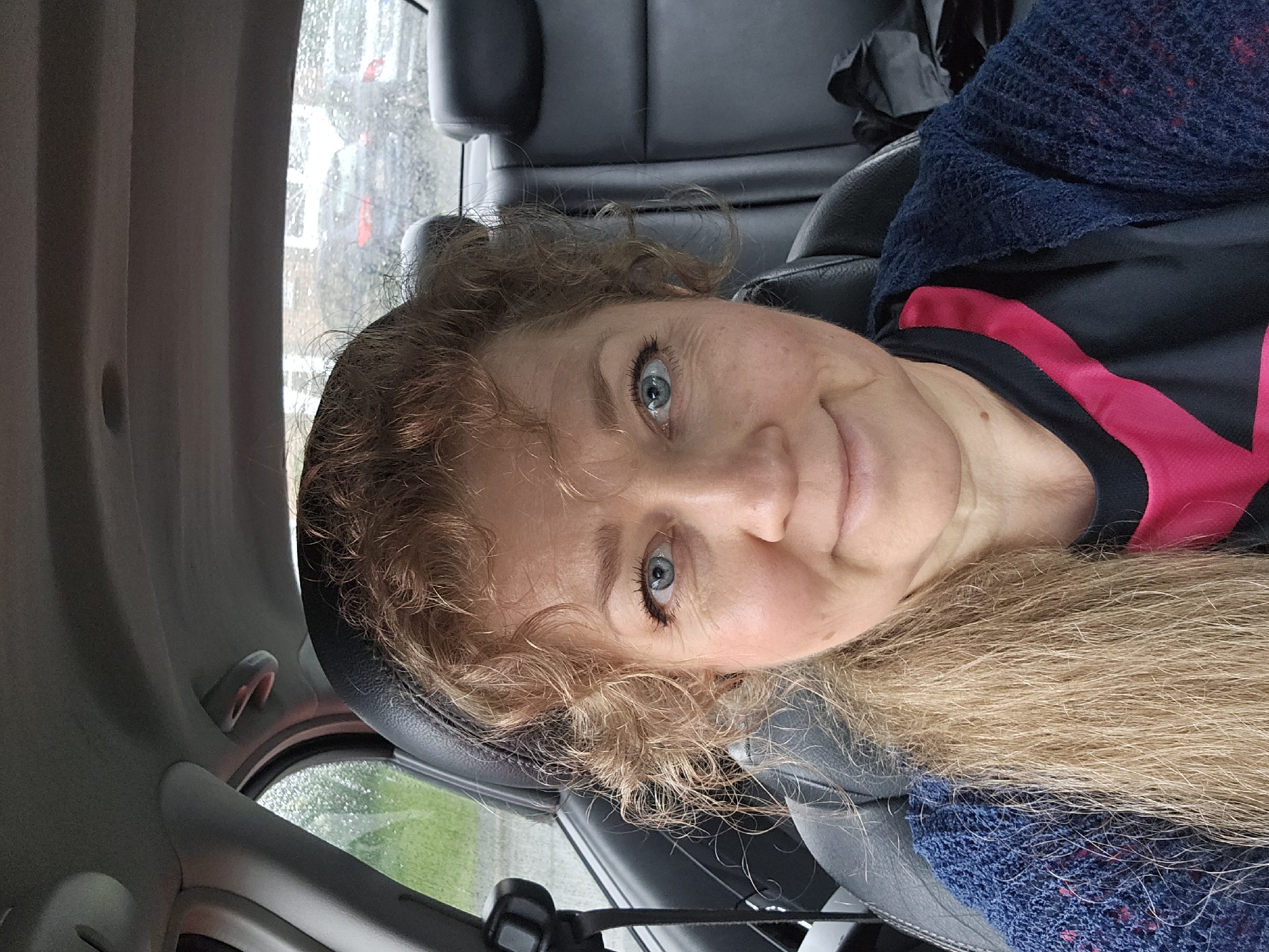 A medium close-up, eye-level selfie shows a woman inside a car, looking towards the viewer with a subtle smile. Her fair skin, bright blue eyes, and light brown/blonde curly hair are prominent. Her hair cascades over her shoulders and around her face. She wears a black top with a distinctive pink stripe and a blue, loosely knitted garment over it.
The setting is the interior of a car, with black leather seats and grey plastic trim visible around her. Through the car window behind her, visible on the left side of the frame, there are reflections and what appears to be raindrops or a wet surface, suggesting rainy or recently rainy weather outside. Blurred greenery and hints of other structures can be seen through the wet window. The overall lighting suggests it is daytime. The woman's expression is calm and pleasant. The location context provided is Philippeville, Belgium. There is no visible text in the image.