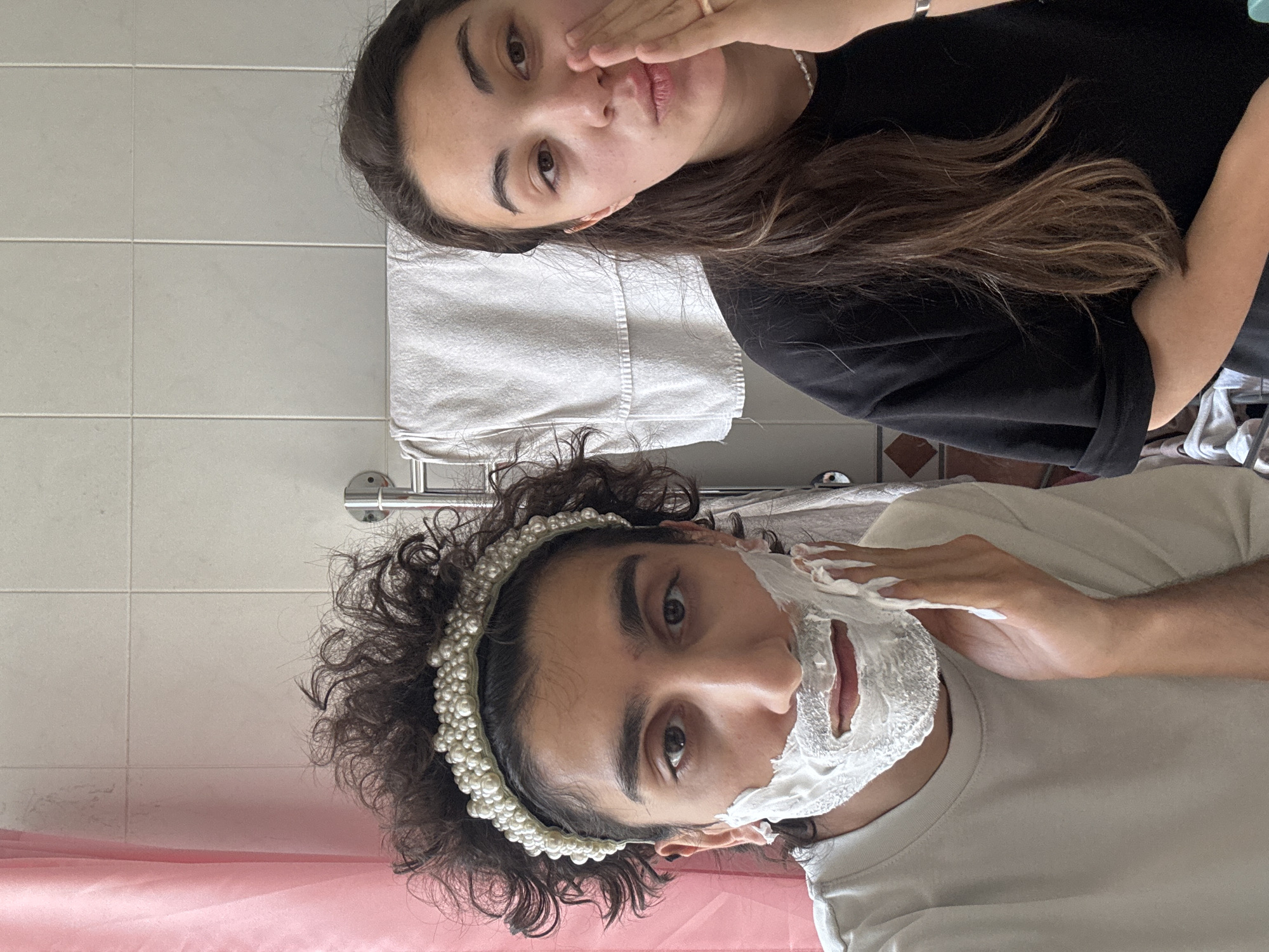 Two individuals are captured in a bathroom setting. One person, positioned in the foreground, is male-presenting with curly hair and a pearl headband, is applying shaving cream to his face. The other person, positioned behind, is female-presenting with long, dark hair and is touching her face with one hand.
The bathroom features white wall tiles and a pink shower curtain. A white towel hangs on a metal rack on the wall behind the female-presenting individual. The specific location within Moscow, Russia is not discernible from the visual information provided.