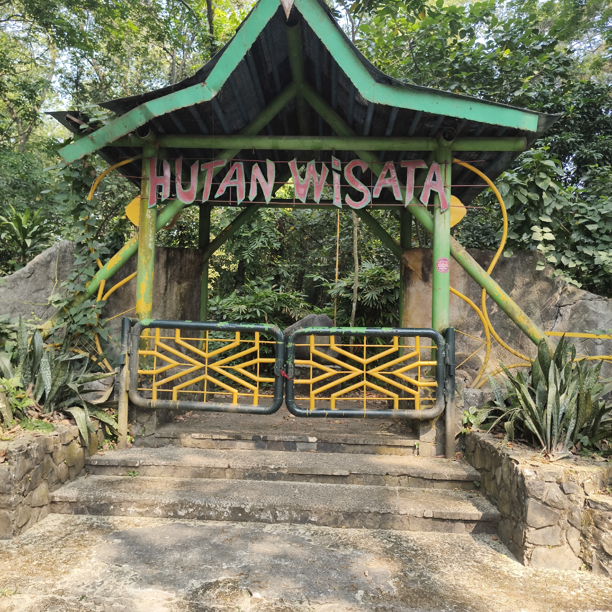 The image shows the entrance to a tourist forest, indicated by the Indonesian text "HUTAN WISATA" which translates to "Tourist Forest". The entrance is marked by a green, pagoda-style roof supported by green and yellow poles. In front of the entrance are two yellow gates with black bars, which are closed. Stone steps lead up to the gates, and the surrounding area is filled with lush green vegetation, including trees and large-leafed plants. The environment appears to be a natural setting, possibly a park or a nature reserve within the city. Based on the style of architecture and vegetation, this entrance likely belongs to Taman Mini Indonesia Indah (TMII), specifically its "Hutan Bambu" (Bamboo Forest) or a similar natural attraction, located in East Jakarta, Indonesia. The scene is bright, with sunlight filtering through the leaves, suggesting it was taken during the day.