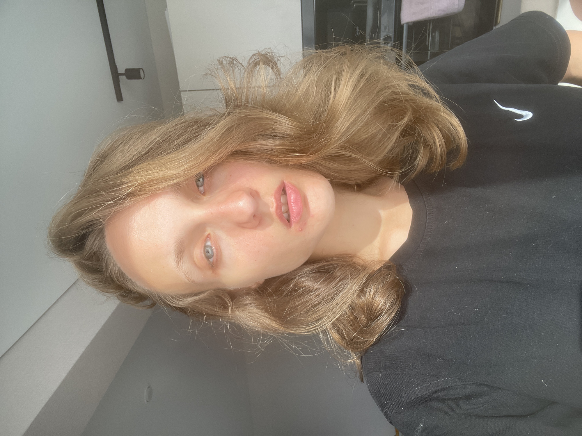 The image shows a young woman with blonde hair and blue eyes. She is wearing a black t-shirt with a white Nike logo. She is looking directly at the camera. The background appears to be an indoor setting, possibly a kitchen, with natural light streaming in from a window. There are some kitchen appliances visible, as well as a light fixture on the ceiling. The woman's skin appears to be slightly flushed, and she has some blemishes on her face. The overall impression is of a casual, candid photograph. The location in the city of Подшивалово, Russia, is not evident from the image itself.