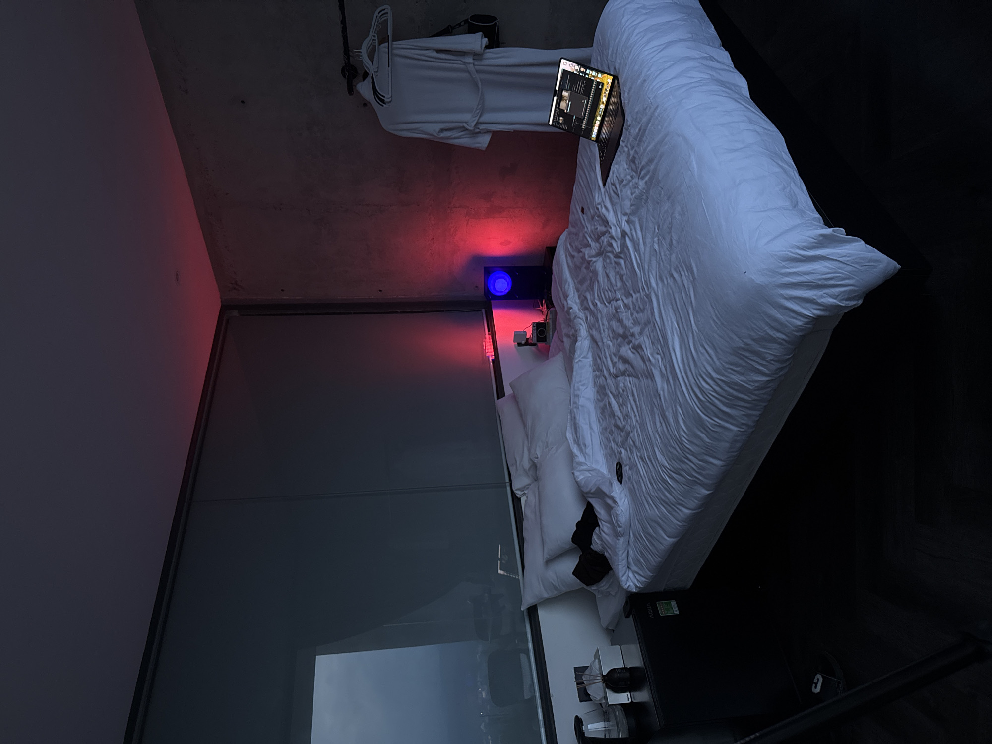 The image depicts a dimly lit hotel room at night. A large bed with white bedding is centrally located. A laptop is open on the bed, displaying a screen. To the left of the bed, a nightstand holds a kettle, tissues, and a diffuser. A bathrobe hangs on a rack on the right side of the room, next to a concrete wall. Red ambient lighting emanates from the ceiling and behind a speaker on the nightstand. The room appears to be located in a high-rise building, suggested by a large window on the left that offers a dark view of a city skyline. There are no people visible in the image. The overall atmosphere is modern and minimalist. The provided context mentions the city of Đa Lộc, Vietnam, however, there are no specific visual cues within the image to confirm this location.