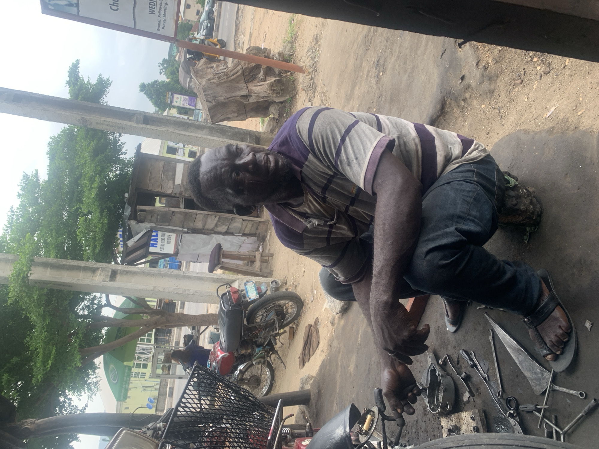 In Lokoja, Nigeria, a dark-skinned man, likely a mechanic, squats on the ground amidst tools and motorcycles. He wears a purple and gray striped polo shirt and blue jeans. His hands are calloused and appear to be resting on his knees. Scattered around him are various tools, including wrenches, pliers, and what appears to be a part of a motorcycle engine. In the background, a red motorcycle is parked, and a woman can be seen in the distance. The scene is outdoors, with a dusty ground and concrete elements, suggesting a workshop or roadside repair area. A large tree provides shade, and a signpost is partially visible. The sky is overcast.