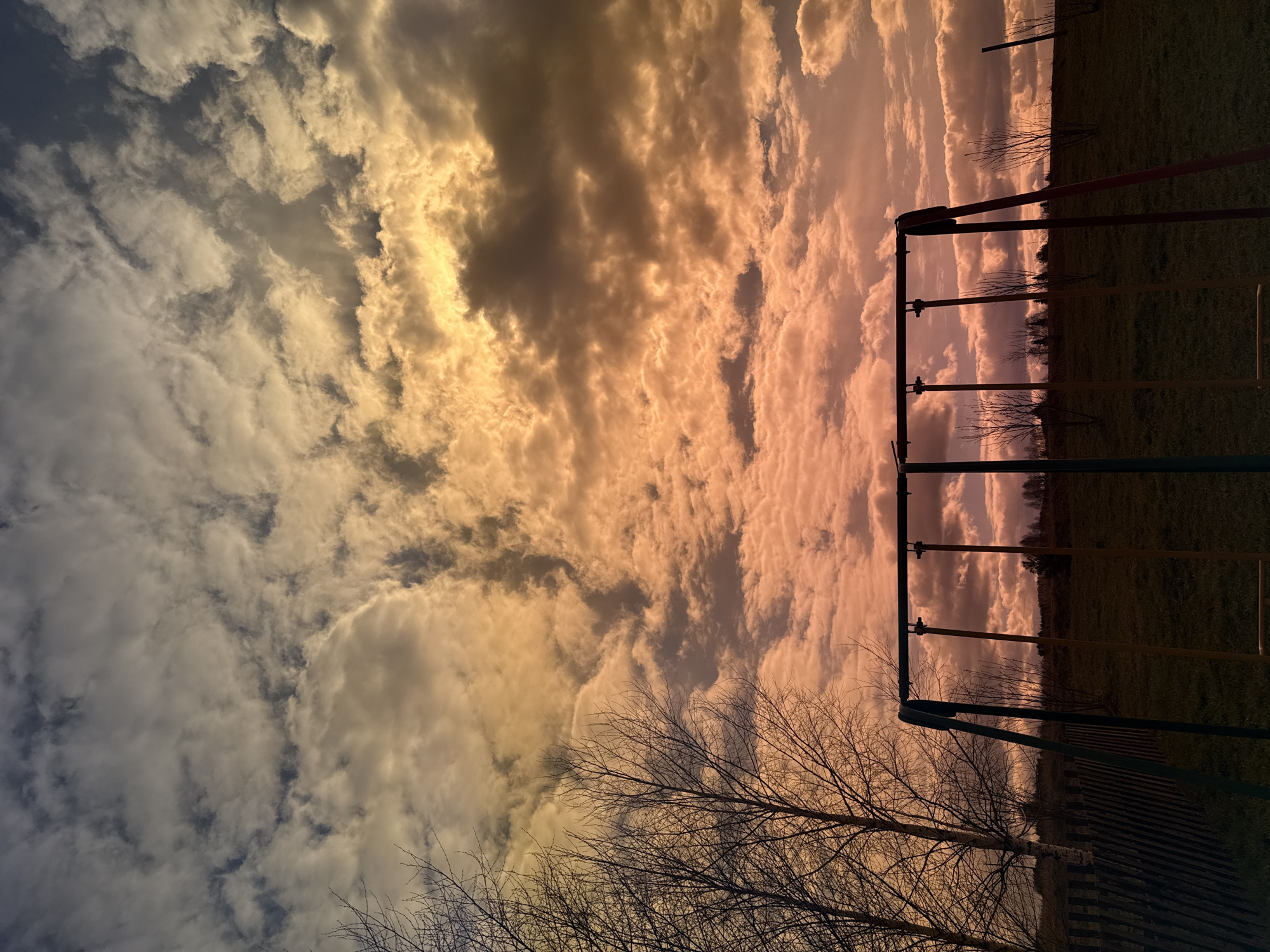 The image shows a view in Nizhnyaya Chermoda, Russia. The sky is filled with clouds transitioning from blue-grey to orange-yellow. A swing set stands on a grassy field. Bare trees are visible in the foreground.