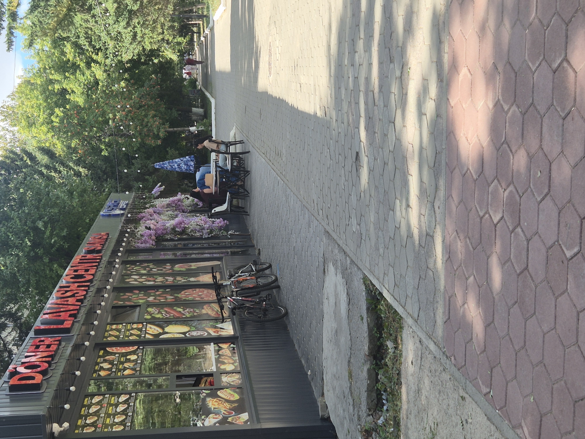 An outdoor scene depicts a food establishment with a prominent sign reading "DONER LAVASH CENTR SAMSA" in red lettering. The establishment features large display windows showcasing images of food items. A string of lightbulbs hangs above the windows, and decorative purple flowers cascade down one side. Two bicycles are parked in front of the establishment. In the background, a paved walkway lined with trees leads into the distance, where several individuals are walking. The ground is paved with hexagonal tiles, with a section of concrete in the foreground. The setting appears to be a park or pedestrian area in Kokshetau, Kazakhstan, likely a commercial zone.