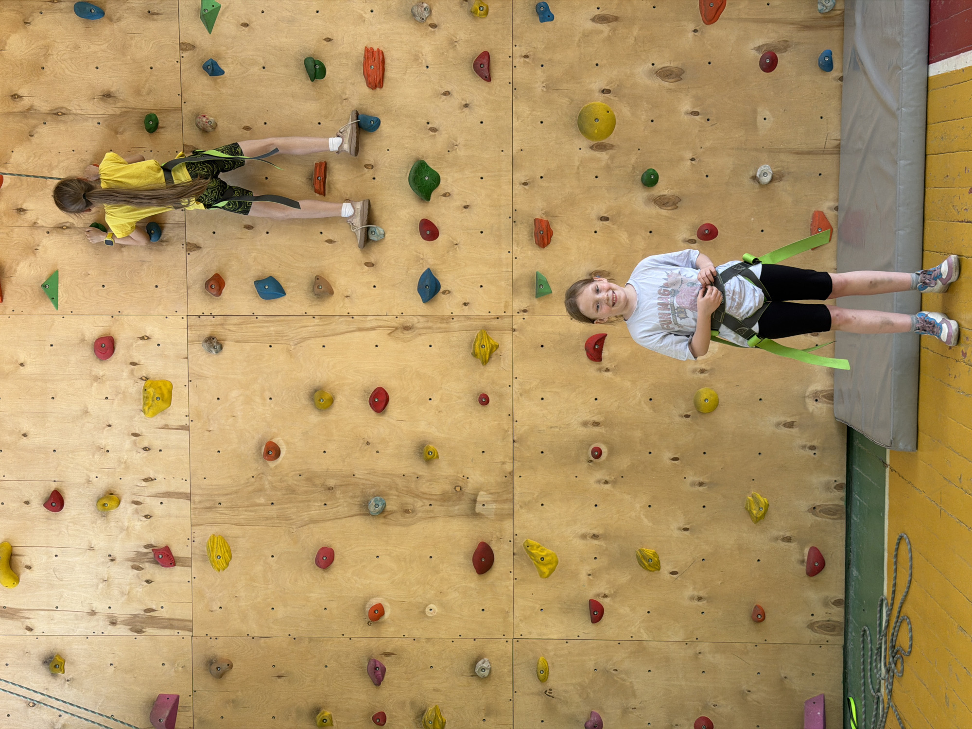 Two girls are positioned horizontally on a plywood climbing wall, presumably in Izhevsk, Russia. The wall features numerous colorful climbing holds of various shapes and sizes. The girl on the left is wearing a yellow shirt and dark shorts, gripping holds with her hands and feet. She has long brown hair. The girl on the right is wearing a light-colored t-shirt and black shorts, and is secured by a climbing harness and a rope. She is smiling and looking towards the camera. A grey crash pad is positioned below the rightmost girl. The wall is made of light wood panels with visible grain. A yellow wall is visible to the right of the climbing wall, with a rope coiled alongside it.