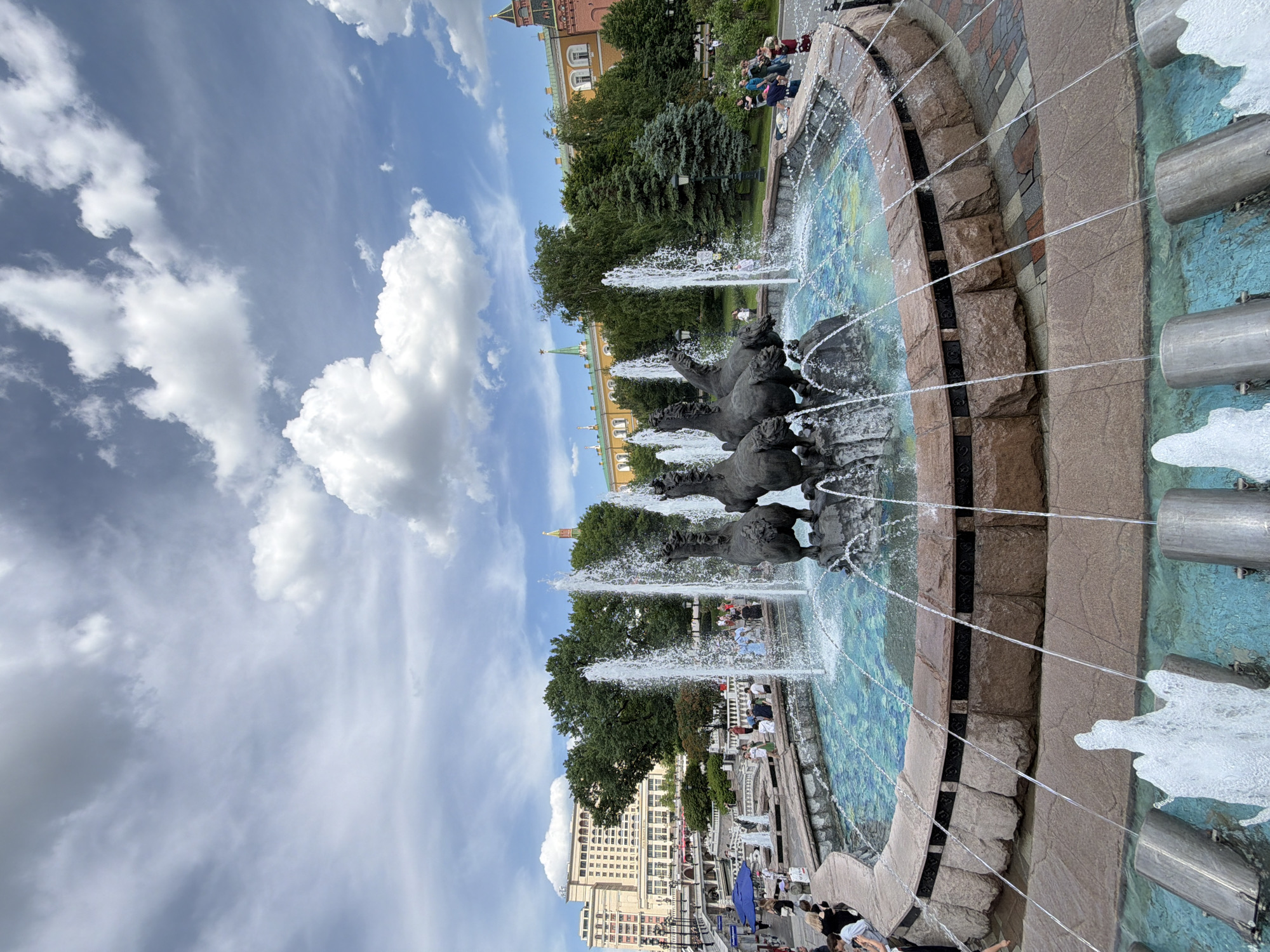 The image captures a vibrant daytime scene of a large, ornate fountain in a public square in Moscow, Russia. The central feature is a circular fountain adorned with multiple dark equestrian statues, depicting horses in dynamic poses, from which water jets spray upwards and outwards into the basin. The fountain's edges are made of rough-hewn stone, and the basin features intricate blue and green tiling. Several horizontal streams of water also shoot from metallic pipes along the outer rim of the fountain.
Numerous people are visible around the fountain and in the surrounding area, enjoying the pleasant weather. Some are casually strolling, while others are seated on benches or ledges under the shade of mature green trees. In the background, distinctive historical buildings with yellow and red-brick facades, peaked roofs, and golden spires or domes are partially visible through the foliage, typical of Moscow's architecture.
The sky is bright blue with scattered white, fluffy cumulus clouds, indicating a clear, sunny, and pleasant day. The overall atmosphere appears relaxed and leisurely, suggesting people are enjoying an outdoor leisure activity. There is no visible text in the image.