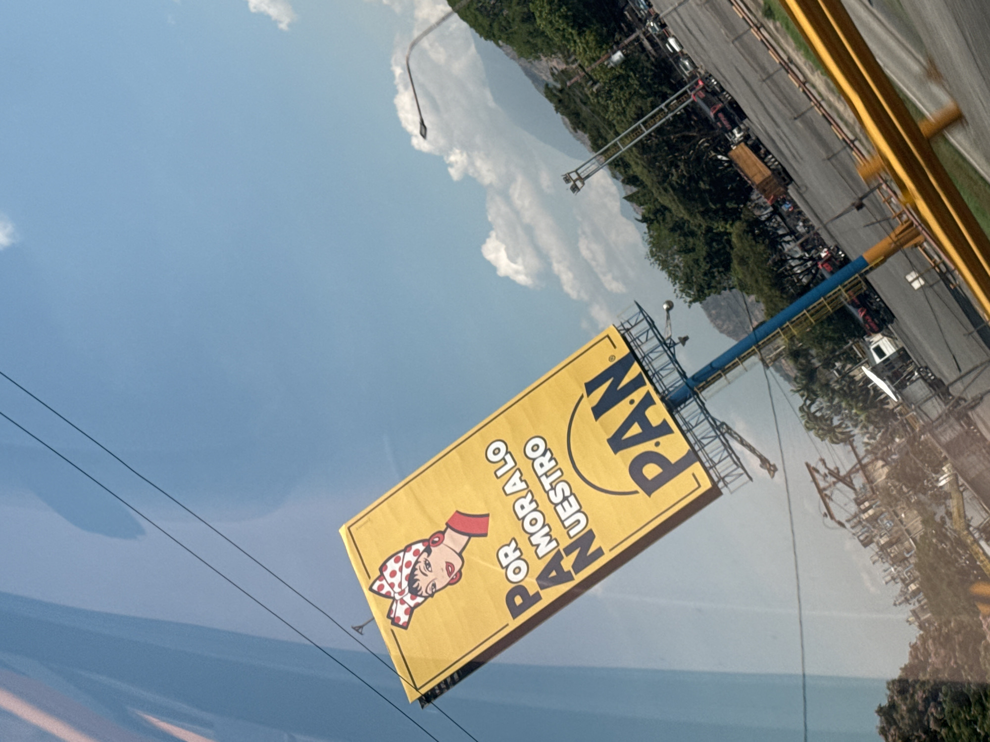 A billboard advertising "Pan Nuestro," or "Our Bread," is visible in Turmero, Venezuela. The sign is yellow with black lettering and features a graphic of a woman wearing a red and white polka dot scarf. The billboard is attached to a metal frame structure. Overhead power lines run in front of the billboard. In the background is a highway lined with trucks and cars. Trees and mountainous terrain are in the far distance. The sky is blue with some clouds.
