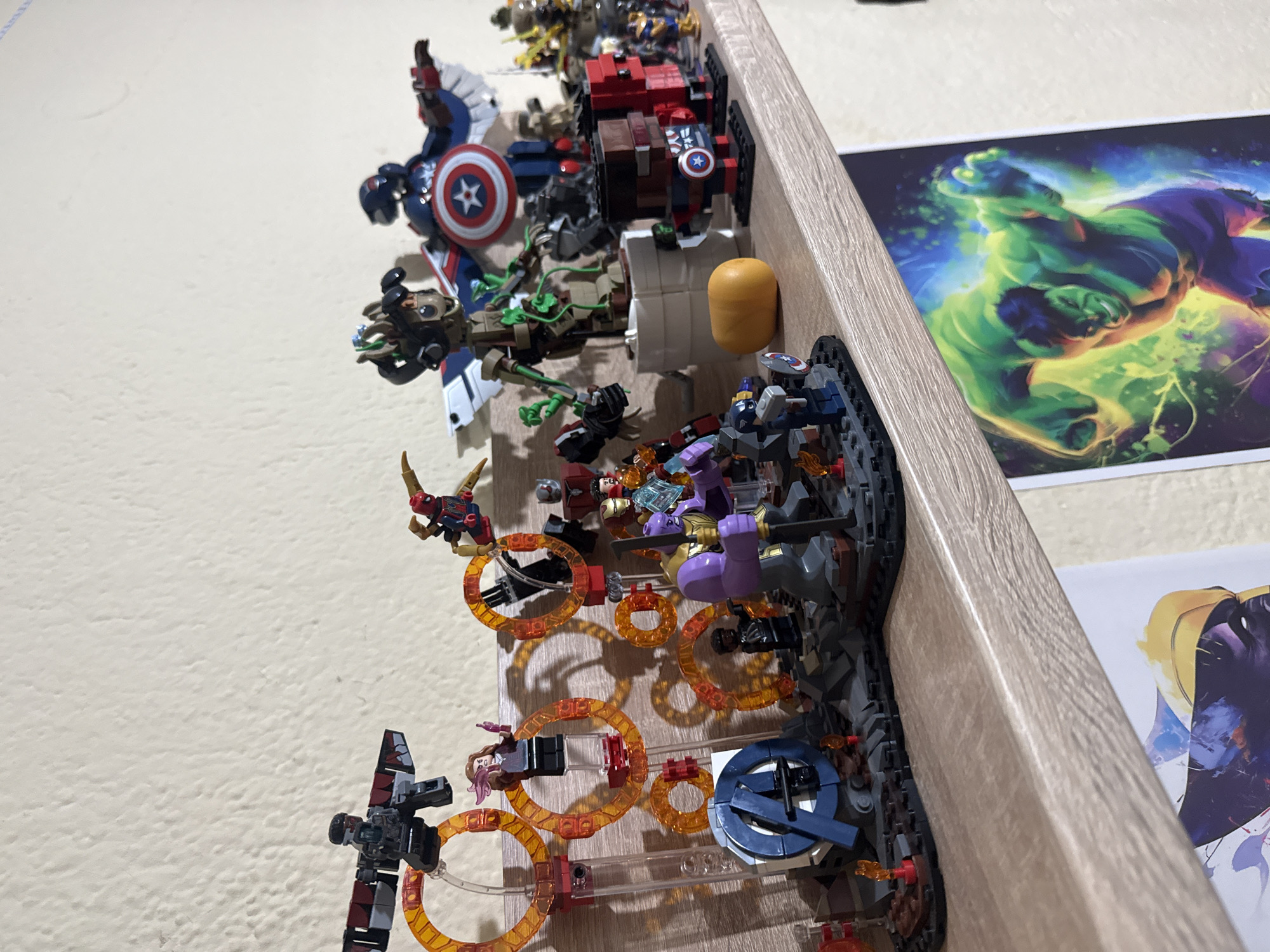 A high-angle, close-up shot shows a shelf cluttered with Marvel Lego figures and associated accessories. Figures include Captain America, Groot, and Thanos. Translucent orange rings and other small pieces are scattered around the figures. A poster of the Hulk and another partially visible poster are positioned behind the Legos. The background wall is off-white. The shelf has a light-colored wood grain. The location is not discernible from the image.