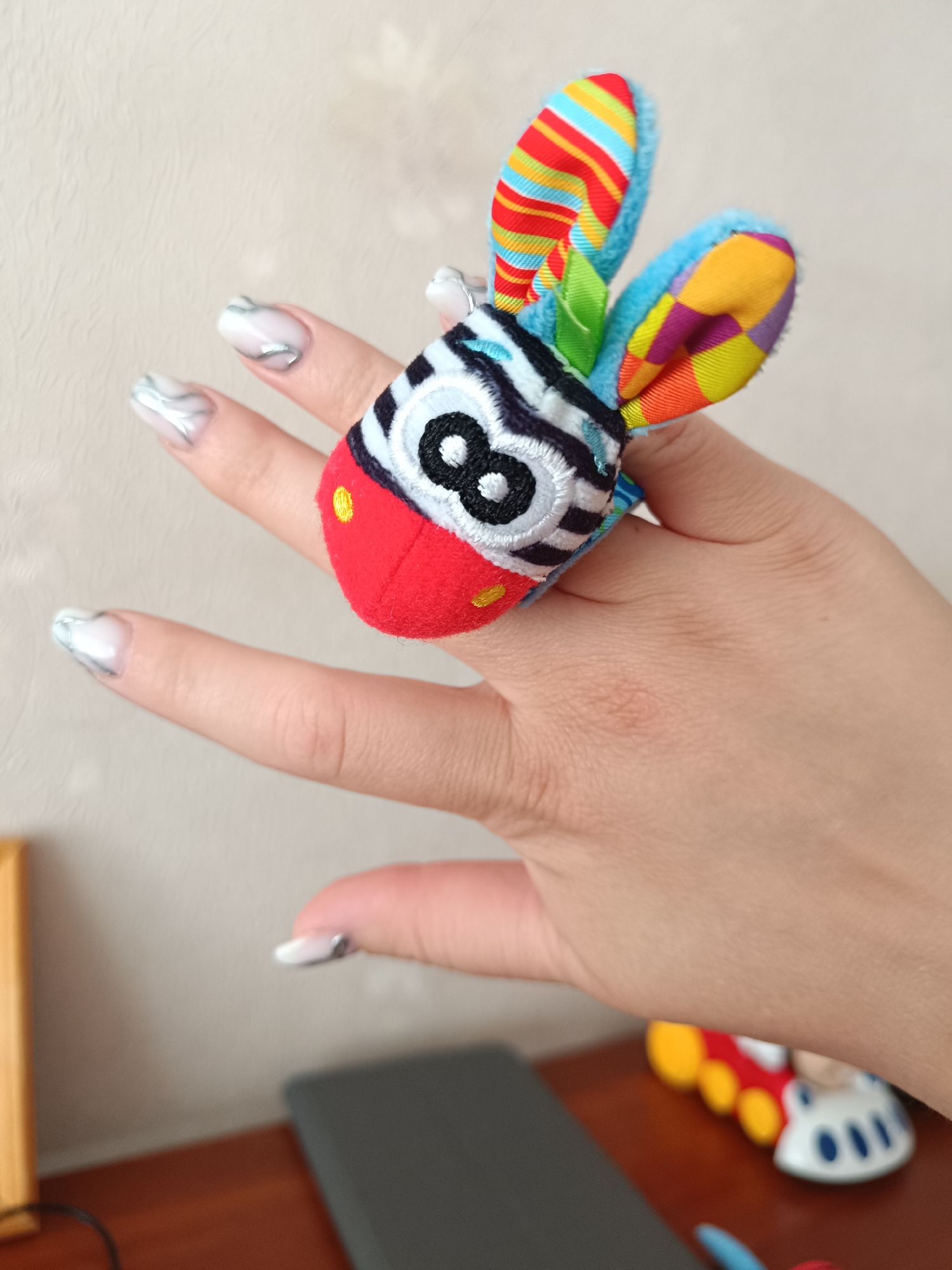 A human hand is presented against a light-beige wall. The fingers are adorned with white and silver marbled nail polish. A colorful, plush toy ring, resembling a zebra with striped ears and a red snout, is worn on the ring finger. A brown table with a gray tablet and a toy car are visible in the background.