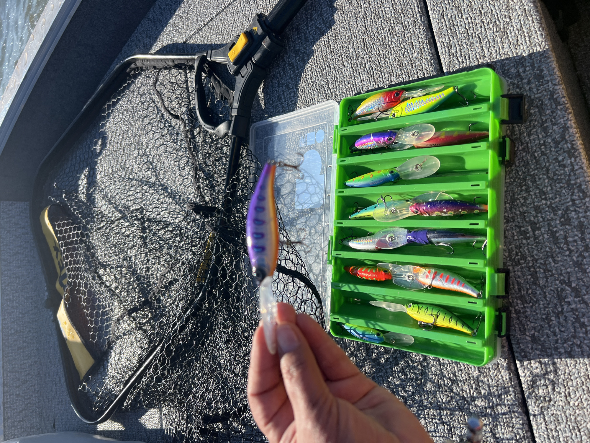 An overhead shot captures a fishing net spread on the floor of a boat. Next to the net, a person's hand holds a purple and orange fishing lure. In front of the net and lure, a green tackle box is open, revealing a collection of colorful fishing lures. The boat's interior is visible, and the water is seen in the background. The image does not provide any information about the city of Novopichugovo, Russia.