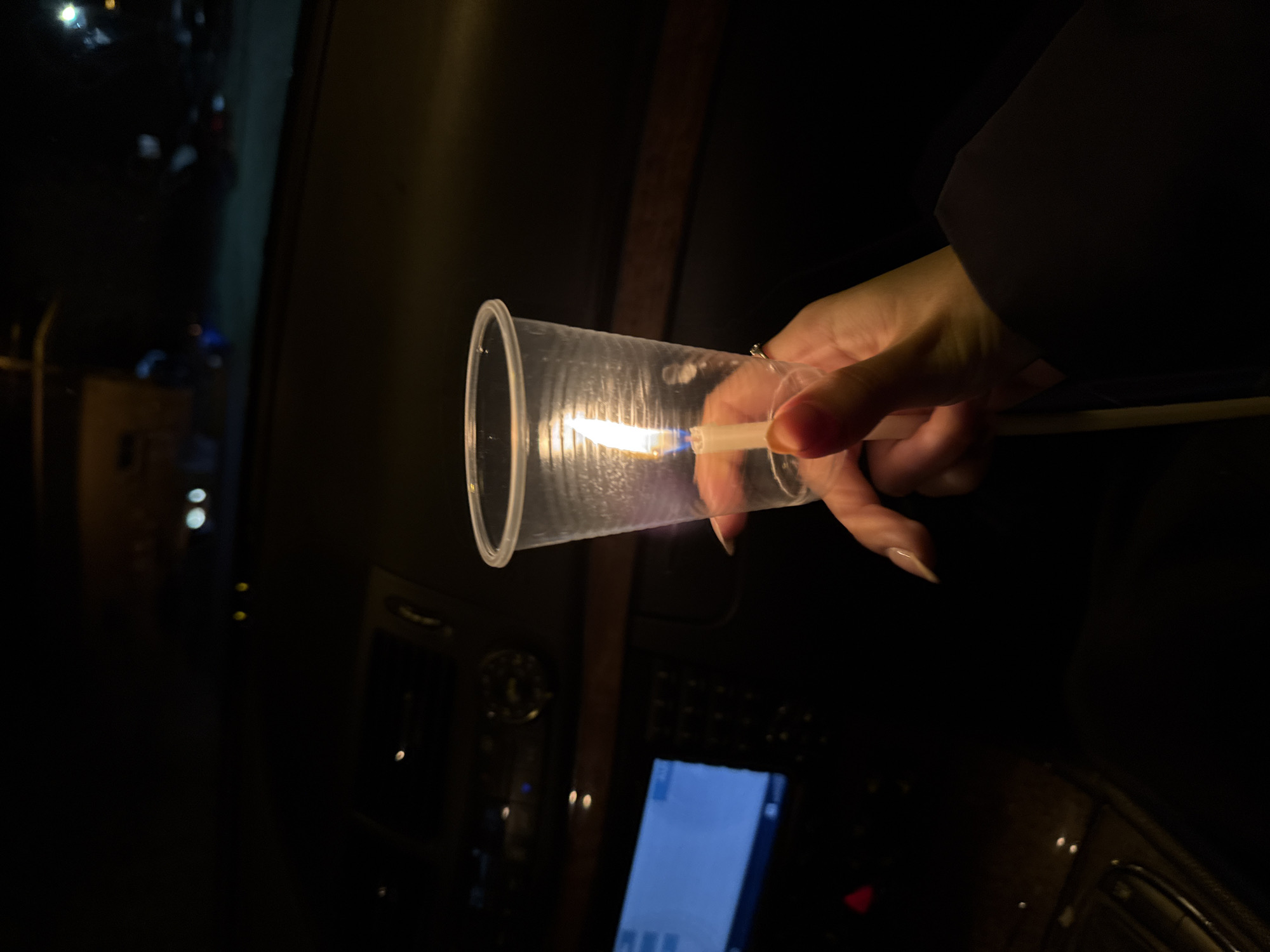 A hand is holding a disposable plastic cup with a lit match inside. The cup is translucent, and the flame is visible within. The background is dark, with the interior of a car visible, including a dashboard with buttons and a screen. The setting appears to be Buhovo, Bulgaria, based on contextual information not directly visible in the image.