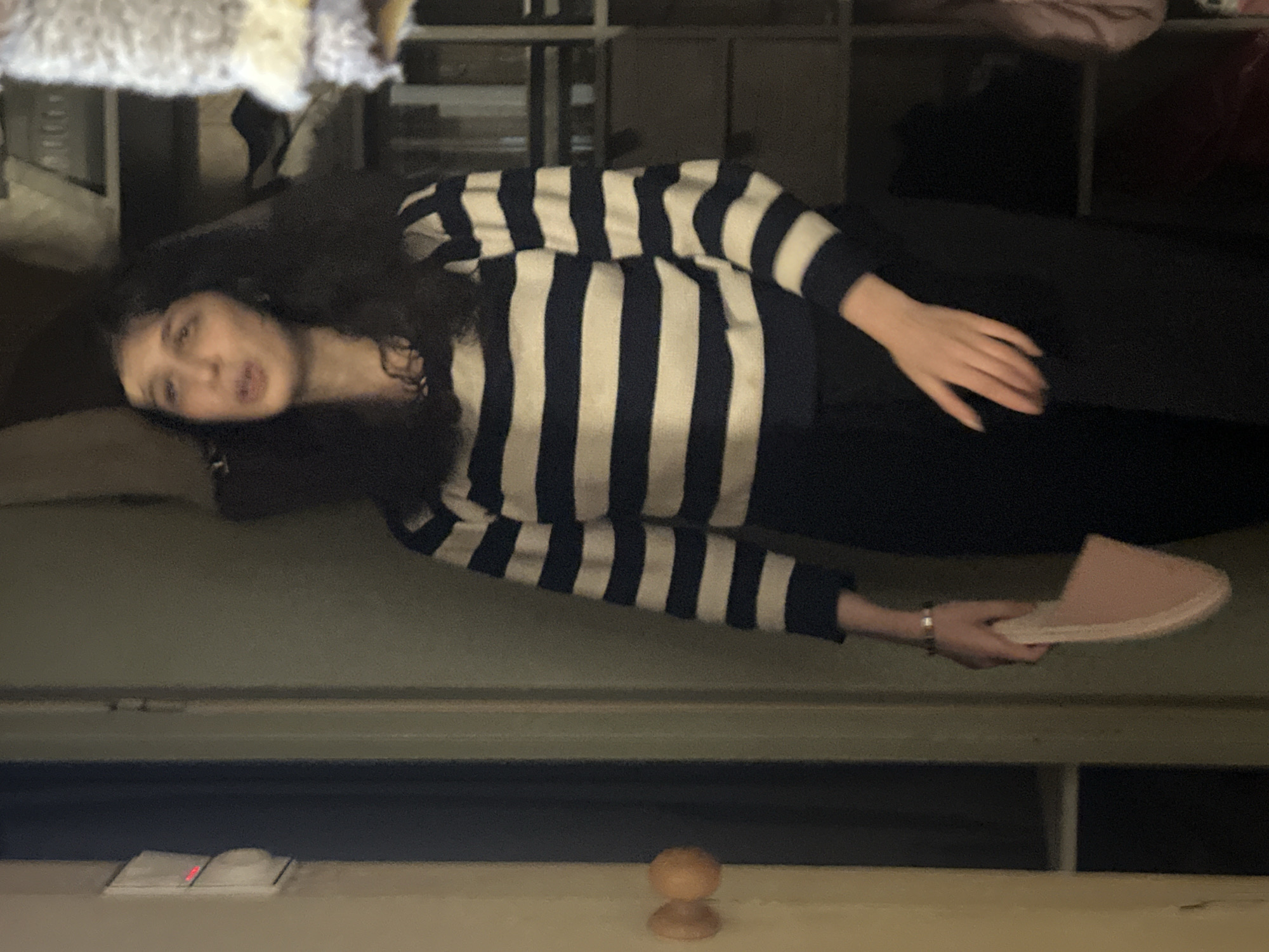 A woman with dark, curly hair is lying on a couch, wearing a navy and white striped long-sleeved shirt and black pants. She holds a pink slipper in her right hand. The location is unidentified, and there is no indication of being in Moscow, Russia. The background includes a closet with items inside and a light-colored surface with a light switch and a small wooden knob.