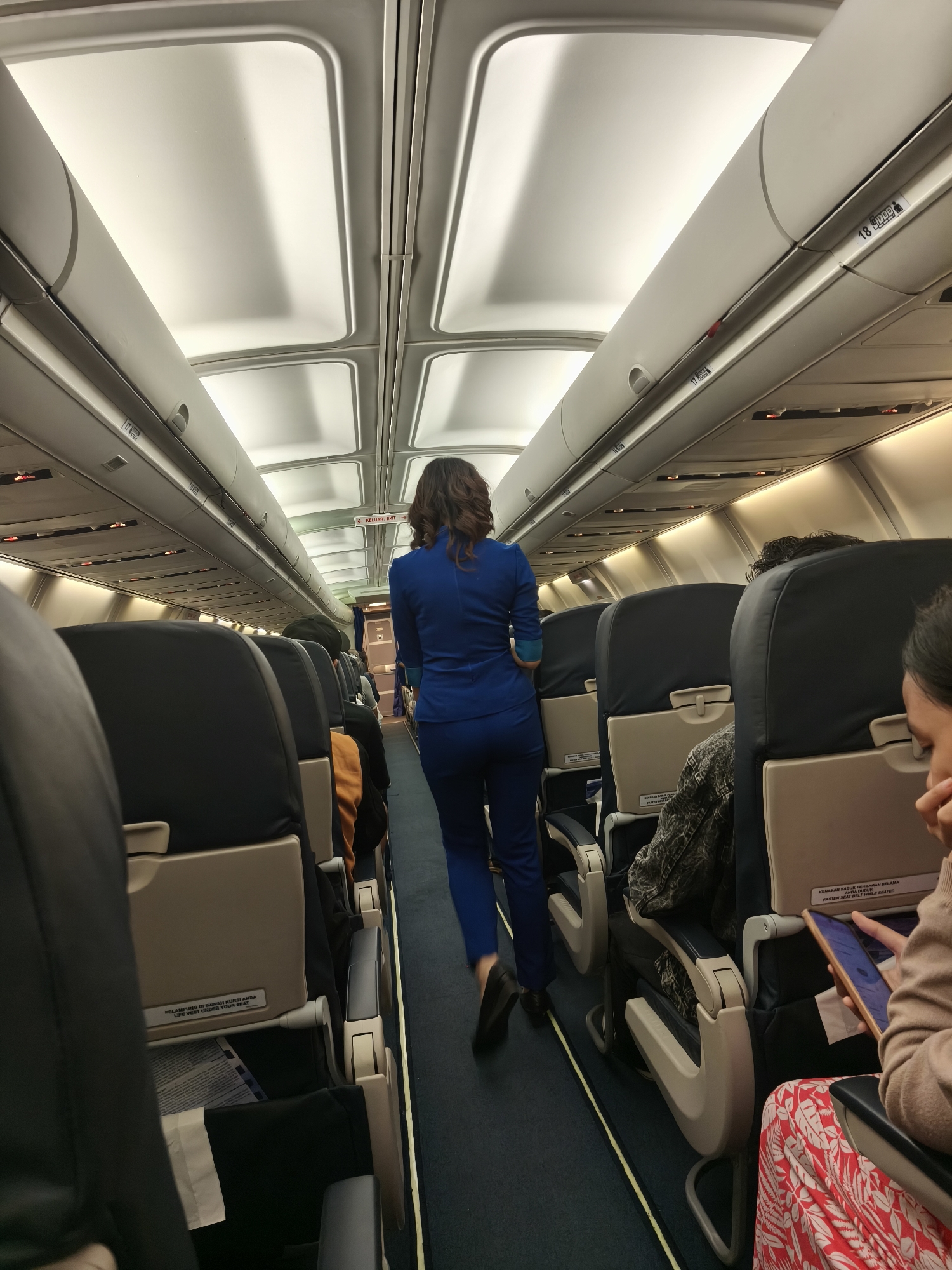 An aerial view of passengers seated inside an airplane cabin with a flight attendant walking down the aisle. The flight attendant is a woman with brown hair, wearing a blue uniform. The passengers are seated in dark blue seats with beige headrests. The cabin is lit by overhead lights. The image shows the interior of an airplane, likely in transit. The location of Kuta, Indonesia is not discernible from the image.