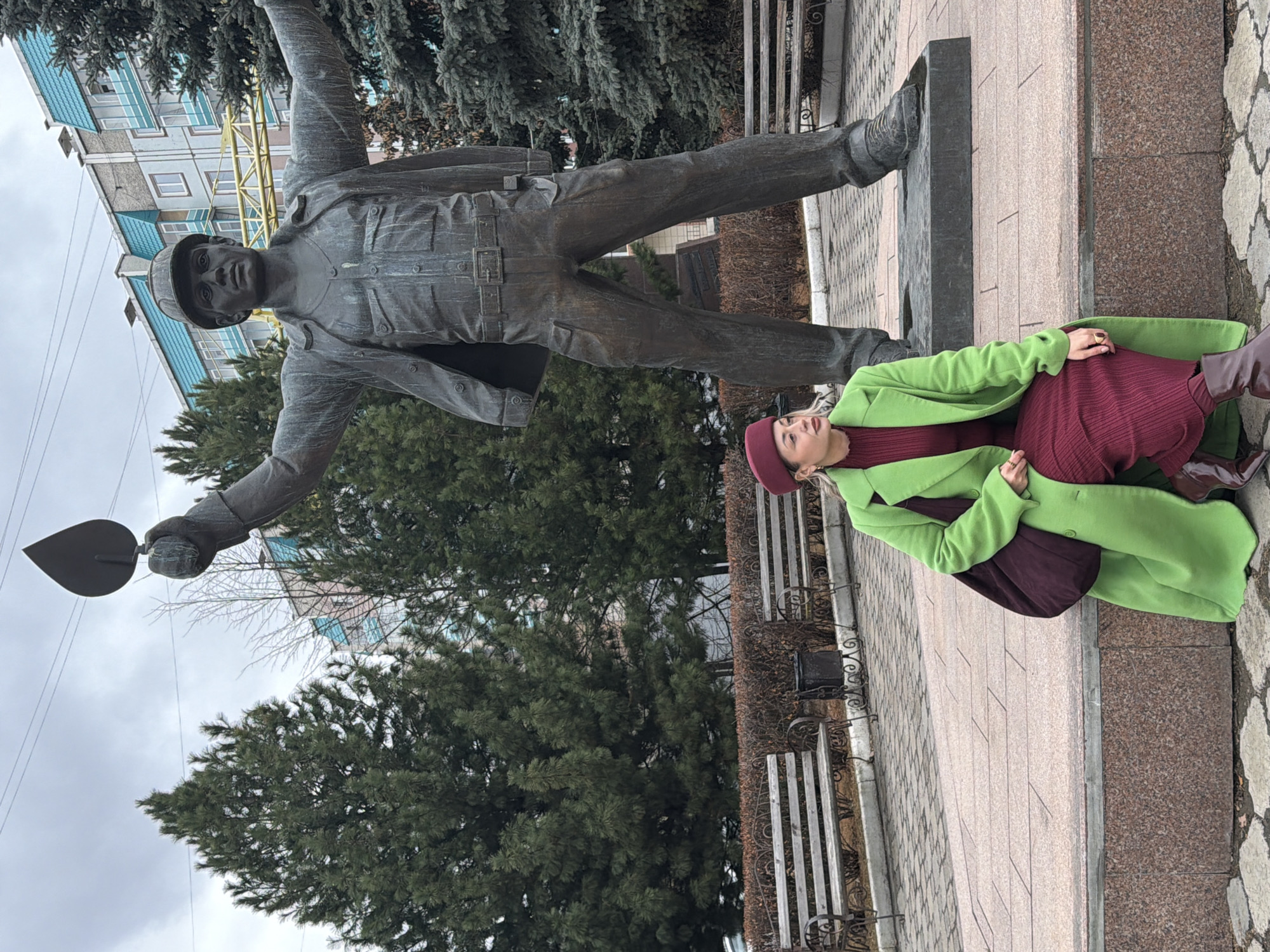 A woman wearing a green coat, burgundy dress, and matching beret lies on a stone ledge next to a bronze statue of a worker holding a trowel. The statue stands atop a tall stone base, and behind it are trees and a building with a blue and white facade. To the side of the ledge is a walkway with benches. The statue is the Monument to the Builders of Krasnoyarsk, located in Krasnoyarsk, Russia.
