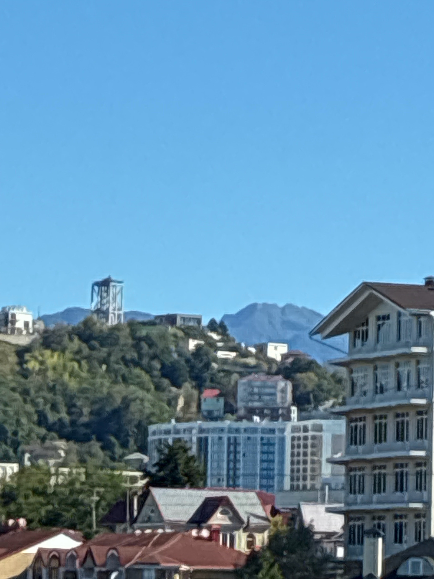 This is a daylight, medium-range, full-shot photograph of a city skyline. In the foreground, several buildings with reddish-brown roofs are visible, some with archways. Behind these, a modern, multi-story white building with many windows is prominently featured on the right. Further into the middle ground, more buildings are present, including a light blue, glass-fronted building. On a hill in the background, a distinctive white tower structure stands against a backdrop of green trees and mountains under a clear blue sky. The scene depicts an urban environment on a hillside, likely in Sochi, Russia, given the architectural style and mountainous terrain. There are no people or animals visible in the frame.