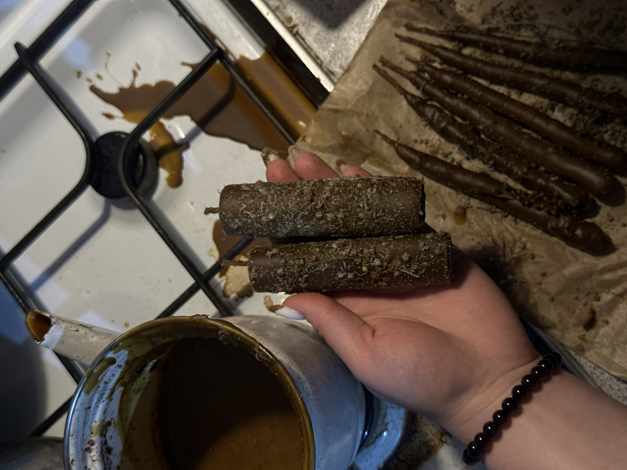 The image shows a person's hand holding two cylindrical chocolate-covered objects that are coated in what appears to be shredded coconut. In the background, there are several long, thin chocolate-covered objects on a sheet of parchment paper. Beside the hand is a white and brown pitcher filled with a brown liquid, possibly chocolate or caramel. A portion of a stove top with brown splatters is visible in the background. The person is wearing a dark red bead bracelet on their left wrist. It is not possible to determine the location of the image or any information regarding the interactions and environment.