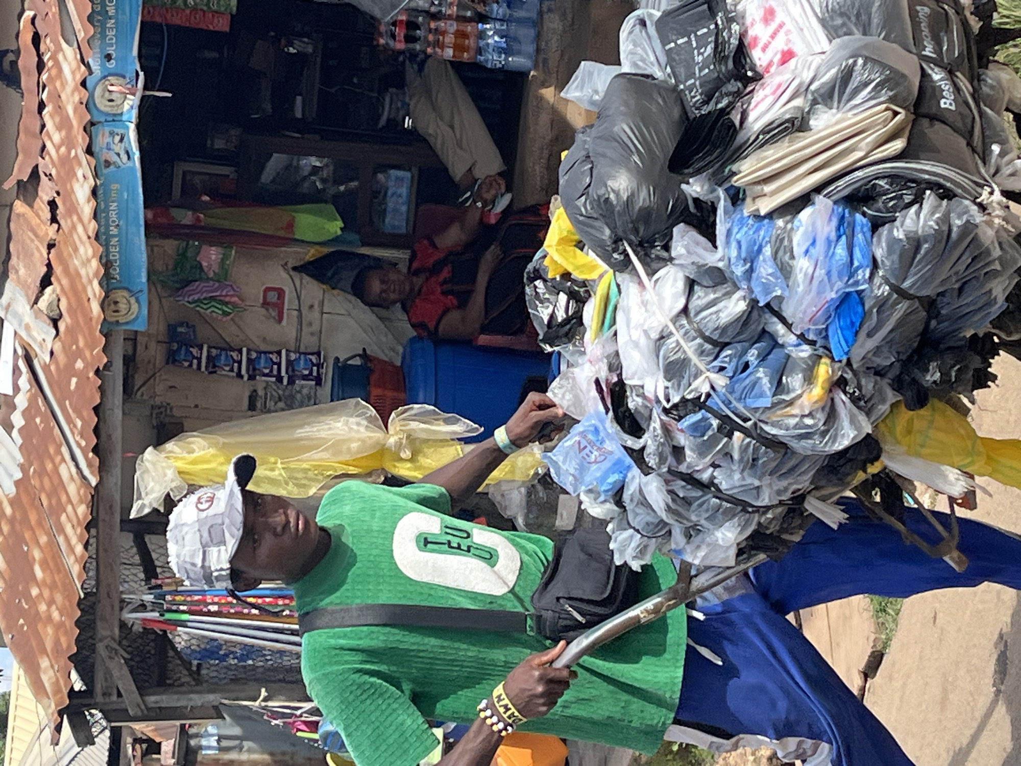 In Jos, Nigeria, a young man wearing a green t-shirt, blue pants, and a patterned cap pushes a cart loaded with bundled plastic bags. He has bracelets on both wrists. In the background, a woman in a red and black dress sits inside a small shop with a corrugated metal roof. The shop displays various items, including drinks and packaged goods. The scene depicts a typical street scene in a Nigerian city, likely a market or a commercial area.
