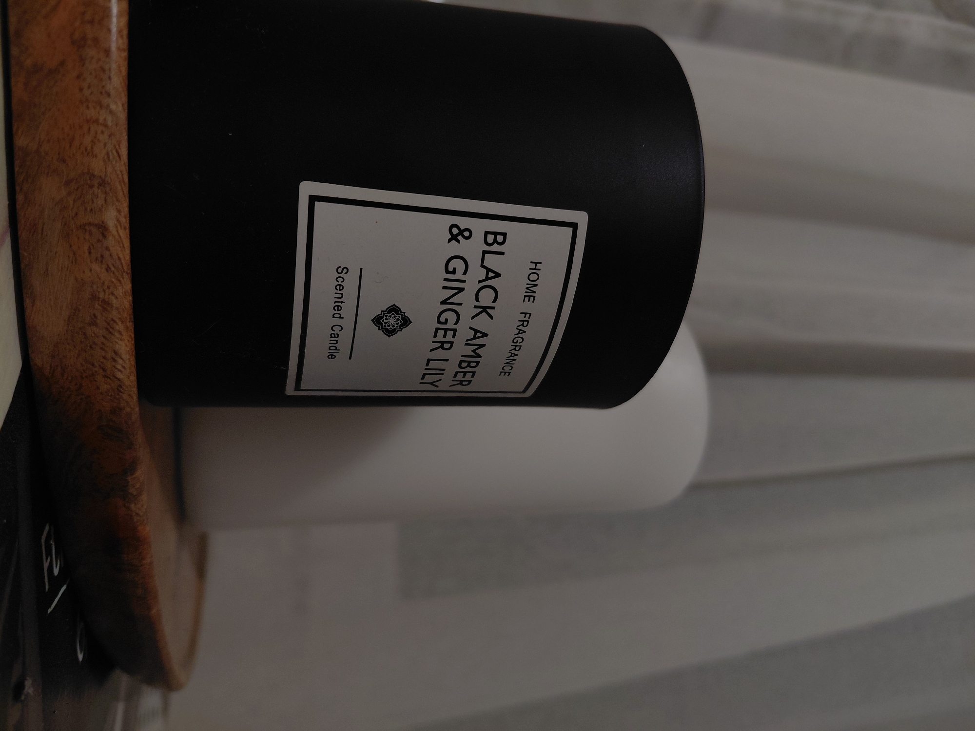 An indoor, eye-level shot focuses on two unlit candles resting on a wooden tray, set against a draped fabric background. A cylindrical, matte black scented candle with a white label reading "HOME FRAGRANCE," "BLACK AMBER & GINGER LILY," and "Scented Candle" is prominently positioned in the left foreground. Behind and to its right, partially obscured, is a similar cylindrical, matte white candle. Both candles rest on a light brown wooden tray with a visible grain. Beneath the tray, a dark, flat surface is visible, possibly part of a book or magazine, showing faint white markings or text. The background consists of light grey or off-white draped fabric, likely curtains, exhibiting soft vertical folds. The lighting is diffused and indirect, creating a subdued atmosphere. There are no individuals present in the image, and no active interactions or actions are depicted. The scene is located in Plovdiv, Bulgaria; however, there are no visually discernible elements within the image itself that specifically identify this geographic location.