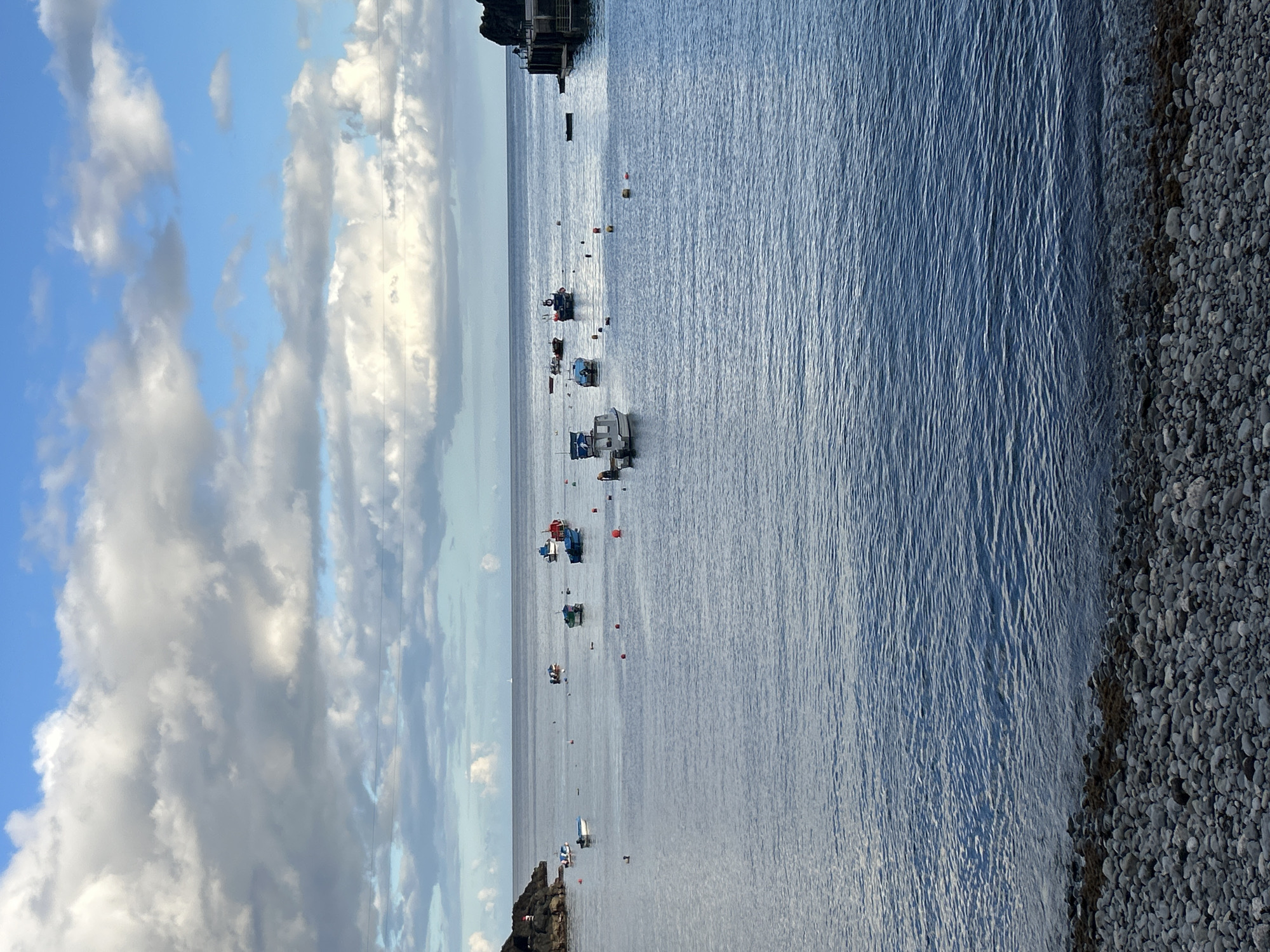The photograph captures a coastal scene in Câmara de Lobos, Portugal. The sky exhibits a blue hue with patches of white cumulus clouds. The sea, stretching out towards the horizon, displays ripples and a darker blue tone. Several small boats are anchored in the water, marked by a series of buoys. A rocky shoreline borders the water's edge on the right. A small cliff topped with buildings is seen in the distance on the top right.