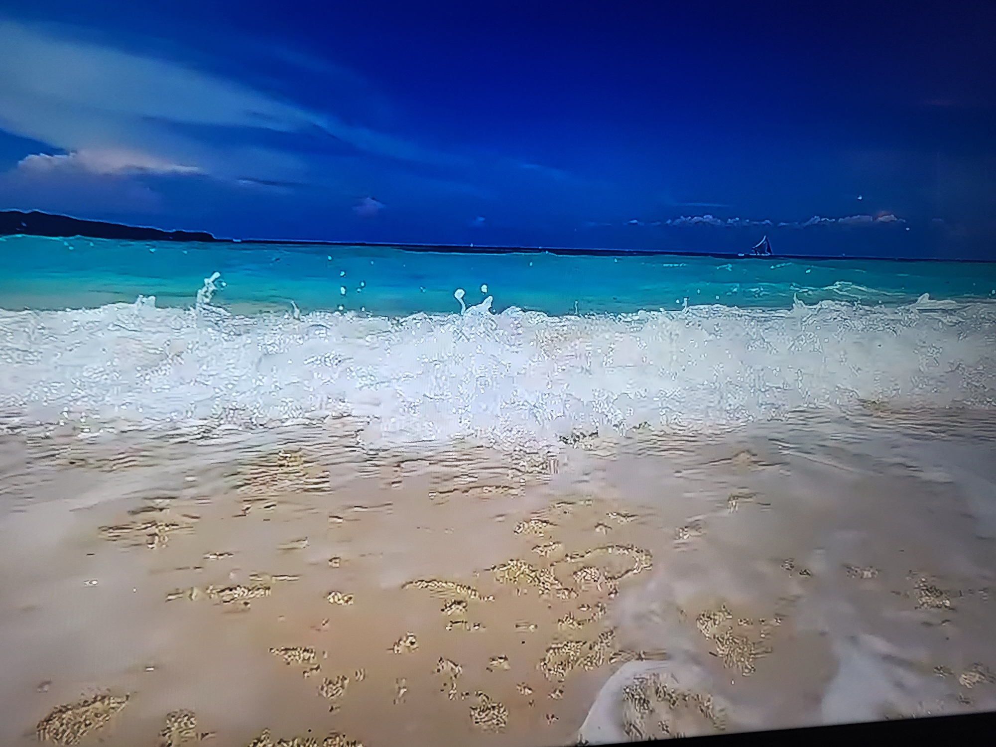 The image shows a serene beach scene with vibrant blue skies and turquoise water. In the distance, a lone sailboat is visible on the horizon. The foreground is dominated by a foamy wave crashing onto the sandy shore, with ripples and wet sand creating a textured surface. The overall impression is one of tranquility and natural beauty. There is no indication that this image is related to the city of Sofia, Bulgaria.