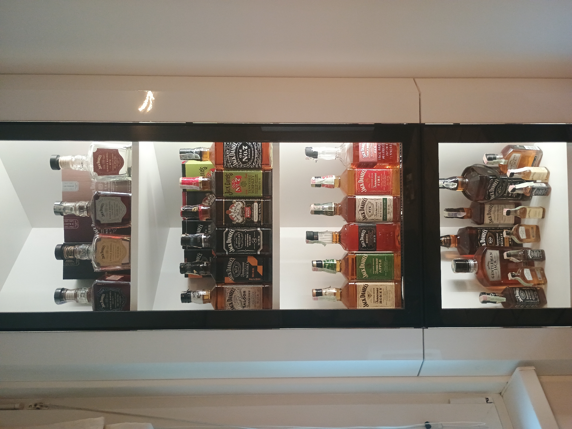 The image shows a display case containing bottles of Jack Daniel's whiskey. There are four distinct sections within the case, each filled with different varieties and sizes of Jack Daniel's bottles. The sections are arranged horizontally, suggesting a collection or presentation of the brand's offerings. The location is unknown.