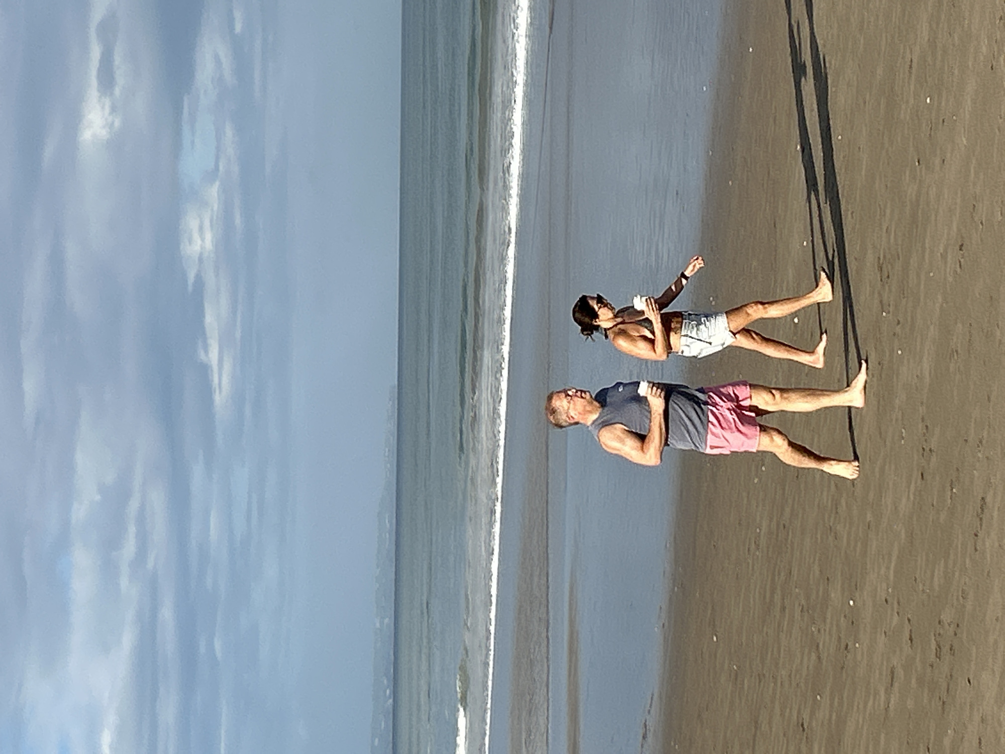 In North Kuta, Indonesia, on a beach, a man and a woman are walking side-by-side on the wet sand near the ocean. The man, wearing a gray tank top and pink shorts, holds a white cup. The woman, in a bikini top and denim shorts, also holds a white cup and wears sunglasses. The ocean is calm with small waves breaking on the shore. The sky is partly cloudy with a hazy horizon visible in the distance.