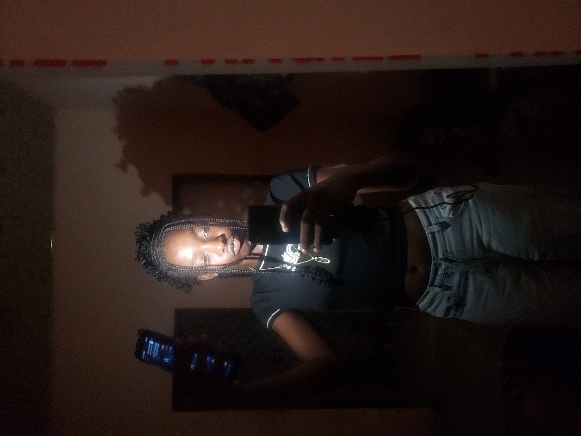 A young dark-skinned individual, likely female, with braided hair, is taking a mirror selfie indoors in the city of Rido, Nigeria. She is wearing a black crop top with white trim on the sleeves, light blue jeans, and a gold chain necklace with a rectangular pendant. The scene is dimly lit, with a distinct light source positioned above her, illuminating her face and casting strong shadows on the wall behind her. In her left hand, she holds a dark-colored smartphone, presumably the camera for the selfie. In her raised right hand, she holds a second dark-colored smartphone displaying a glowing blue screen. The background, visible in the mirror's reflection, consists of a light-colored wall with a dark rectangular object on the left and a dark, irregularly shaped object hanging on the wall on the right. The vertical edge of the mirror's frame is visible on the far right, characterized by white material with intermittent red markings.