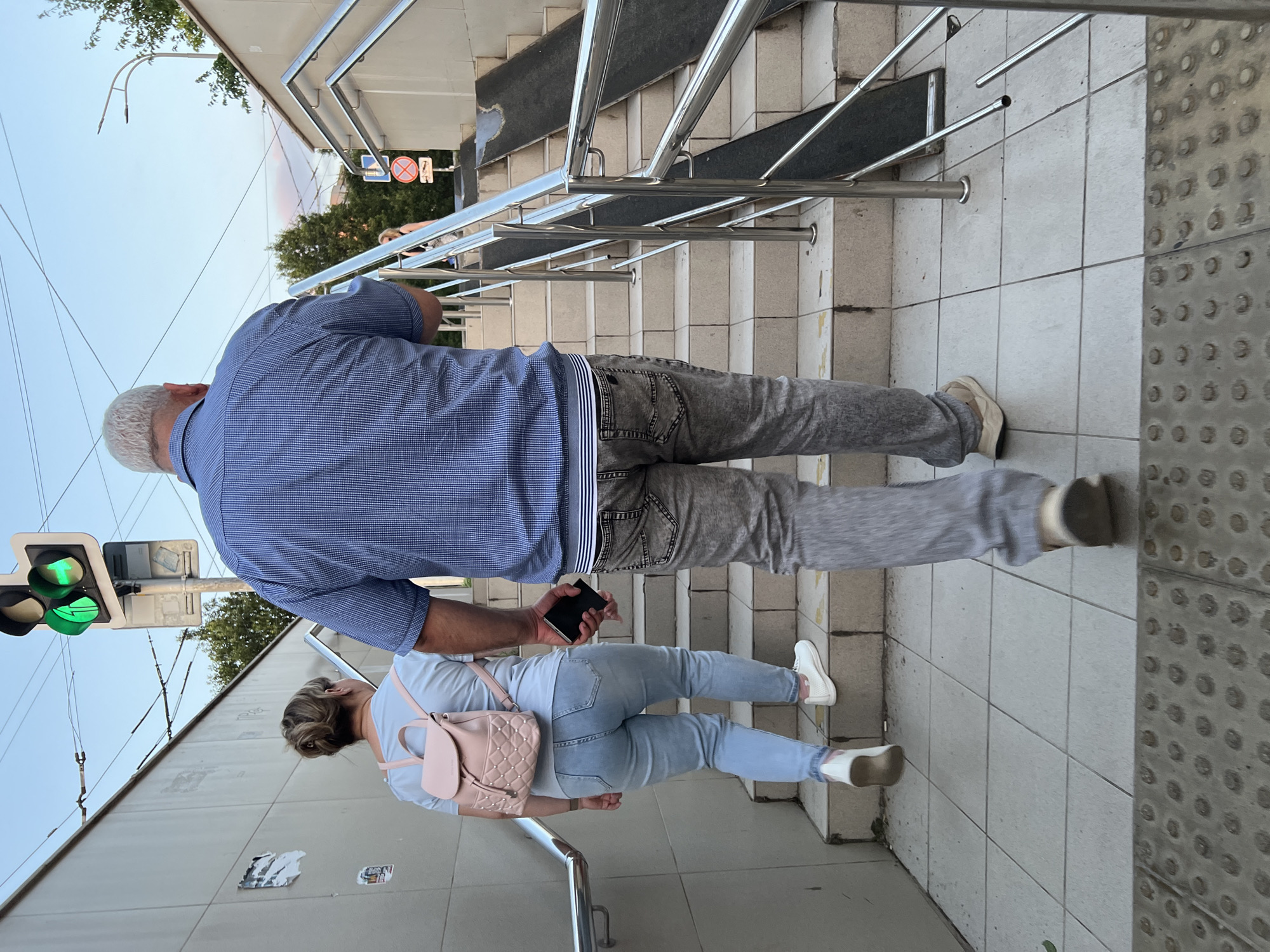 The image captures a street scene in Krasnodar, Russia, featuring a pedestrian underpass with a set of stairs and a ramp. Two individuals, a woman and a man, are ascending the stairs. The woman, in the foreground and to the left, is wearing a light blue top and light blue jeans, carrying a pink backpack. The man, to her right and slightly ahead, is dressed in a blue striped shirt and gray jeans. He is holding a dark object, likely a mobile phone, in his right hand. Above them, a traffic light displays a green signal with an arrow pointing straight ahead. The underpass features tiled walls and stainless steel railings along the stairs and ramp. The overall atmosphere suggests a typical urban environment.