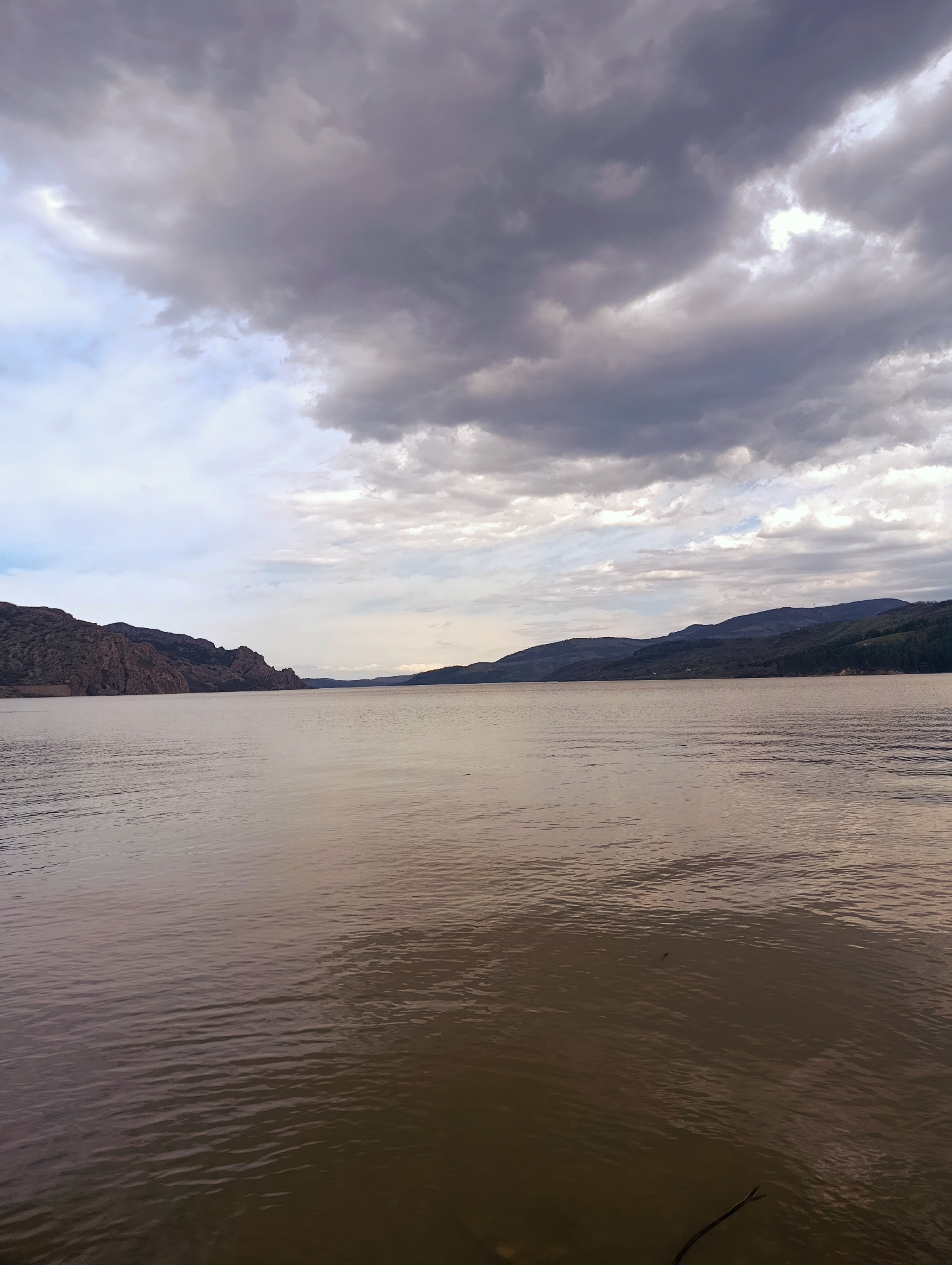 The image shows a body of water, possibly a lake, with mountain ranges on either side. The water is calm with gentle ripples. The sky is overcast with heavy, dark clouds. A thin branch or twig is visible near the bottom right corner of the frame, partially submerged in the water. The location is likely Shiroko pole, Bulgaria, based on the context. The scene depicts a serene, possibly gloomy, landscape. No people or distinct objects are visible on the water or land.