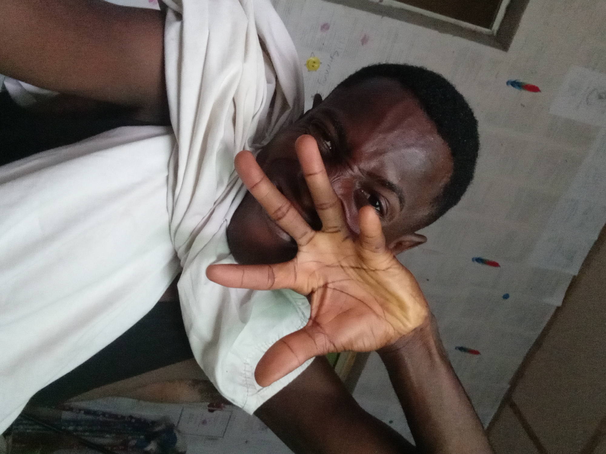 A selfie of a Black male is displayed at an oblique angle. He is wearing a white shirt, and his dark hair is cut short. He is holding up his right hand in front of his face with his fingers spread. His expression is a squinting, slightly pained grimace. The background is a wall or ceiling with some markings or possibly crayon drawings. The location is purportedly Akwete, Nigeria, though no specific visual cues confirm this.