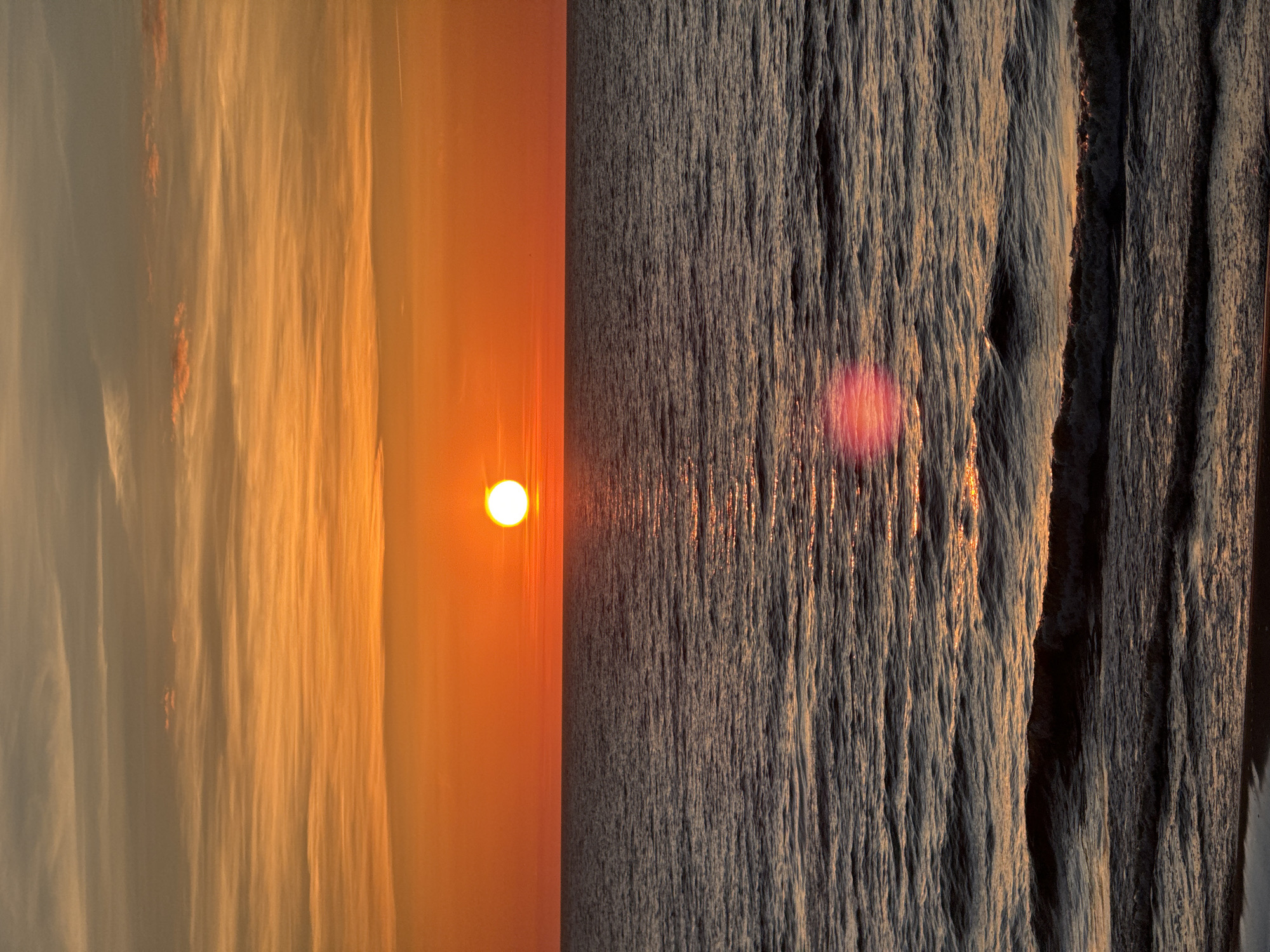 The image depicts a sunset over the Baltic Sea near Garupe, Latvia. The left side features a hazy, bright orange sky with the sun visible as a small, bright orb. The right side shows dark, turbulent waves reflecting the orange sunlight, creating a textured surface. The image is divided vertically, contrasting the smooth sky with the rough sea.