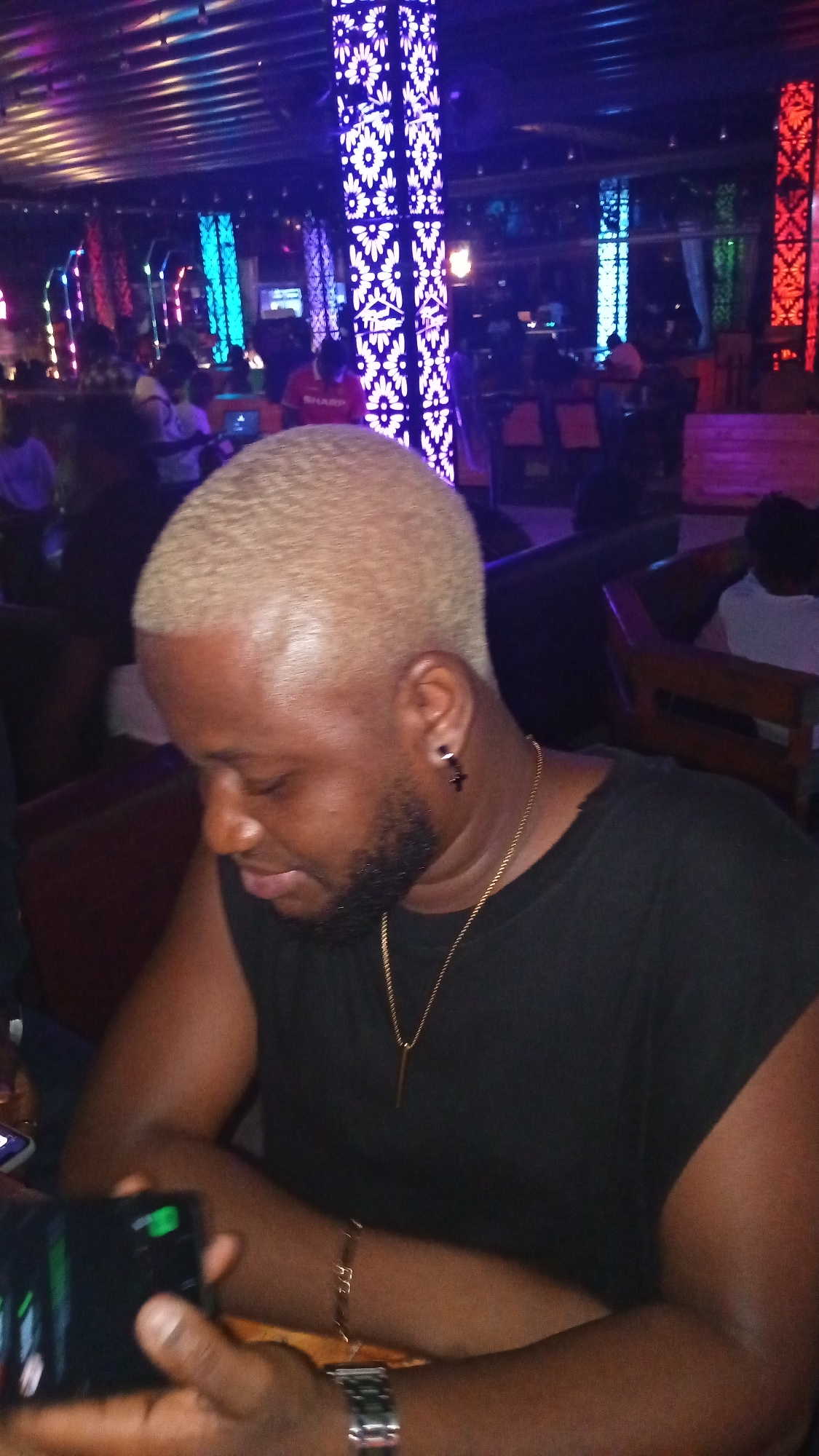 The image shows a close-up of a man with bleached blonde hair and a beard, wearing a black t-shirt, a gold necklace, and a silver watch. He is seated in what appears to be a dimly lit nightclub or bar, with colorful, illuminated pillars in the background. The environment suggests a vibrant, nighttime social setting, typical of entertainment venues in cities like Howayi, Nigeria. The man is looking downwards, possibly at a phone he is holding in his left hand, which is partially visible. The overall atmosphere is relaxed and casual.