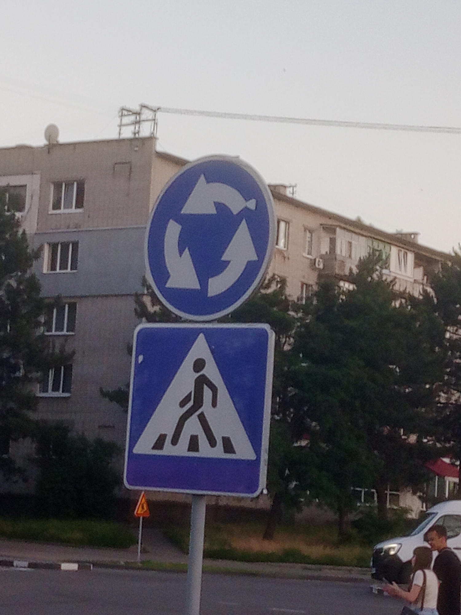 In Horishni Plavni, Ukraine, at street level, two traffic signs are prominently displayed. The upper sign is circular with a blue background and three white arrows indicating a roundabout. Below it is a blue rectangular sign with a white triangular inset containing a black silhouette of a pedestrian crossing. A smaller orange triangular sign with a black symbol is visible in the background to the left. A white van is parked on the right side of the road, with two people, a man and a woman, standing near it. The man is wearing a black t-shirt, and the woman is wearing a white top and blue shorts. They appear to be interacting with each other. In the background, a multi-story apartment building stands behind dense green trees. The sky is overcast and bright.