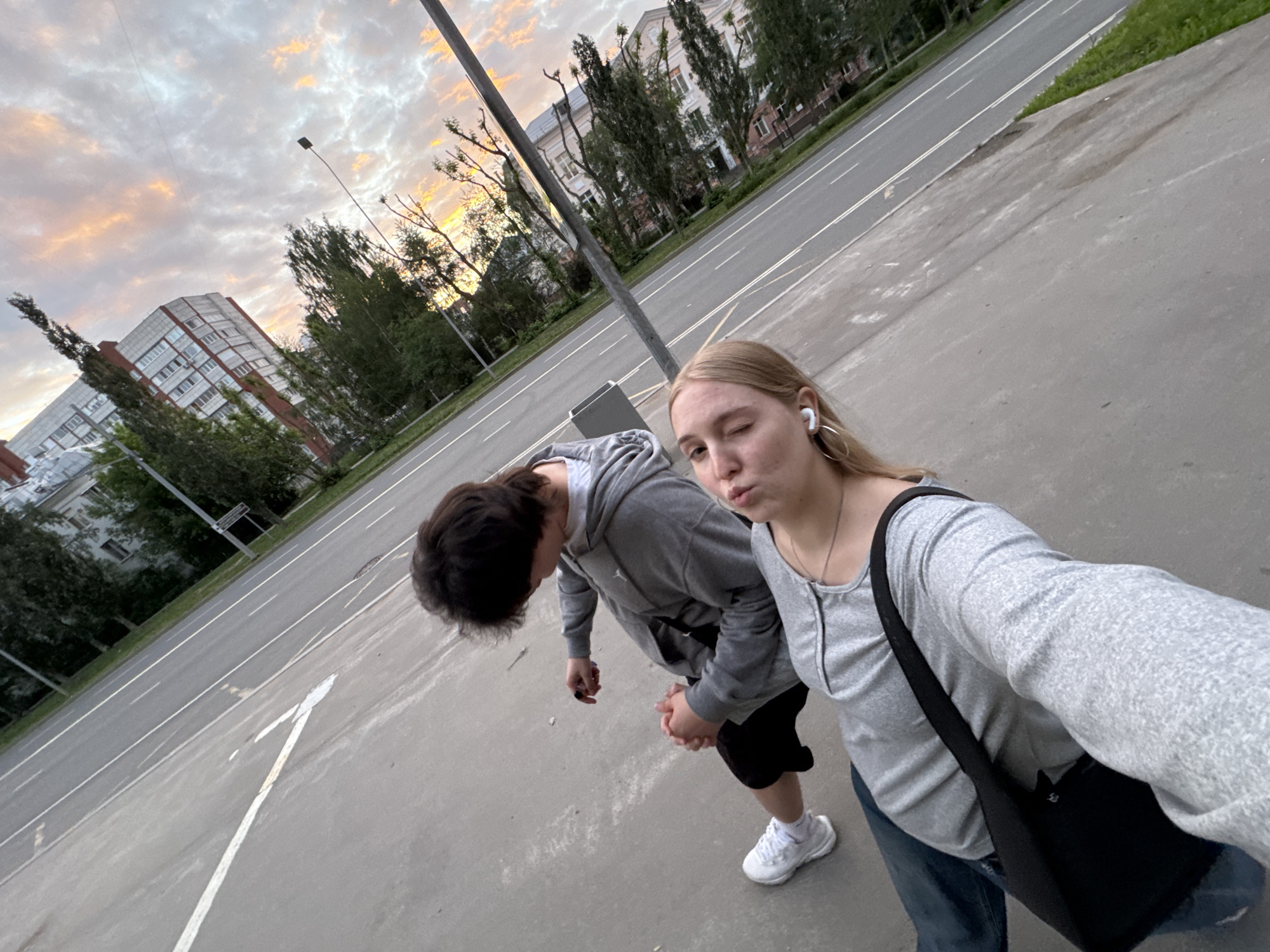 The image presents a tilted, eye-level perspective of an urban scene in Perm, Russia, at dusk or dawn. Two individuals are positioned on a paved surface alongside a road, beneath a sky displaying a mixture of grey clouds and bright orange and pink sunset/sunrise colors.
In the foreground, a female with blonde hair, wearing a light grey long-sleeved top, blue jeans, white wireless earbuds, and large silver hoop earrings, holds the camera with her left hand, suggesting a selfie. She carries a black shoulder bag across her body, and her facial expression is characterized by slightly pursed lips and a partially closed right eye.
Behind and to the left of the female, a male with dark, disheveled hair is observed. He is dressed in a grey hooded sweatshirt, black shorts, and white sneakers. His hands are clasped together in front of him.
The ground beneath the subjects is asphalt pavement, marked with various white lines, including a prominent "Y" shape. To their right, a multi-lane road extends into the distance, delineated by white lane markings. A strip of green grass and numerous trees line the road. In the midground, a dark street light pole stands vertically on the right side of the frame.
The background features urban architecture consistent with Russian cities. On the left, a tall, multi-story residential building with light-colored facades and numerous windows is visible through scattered trees. Further down the road on the right, additional buildings and trees are present, partially illuminated by the sky's warm hues.