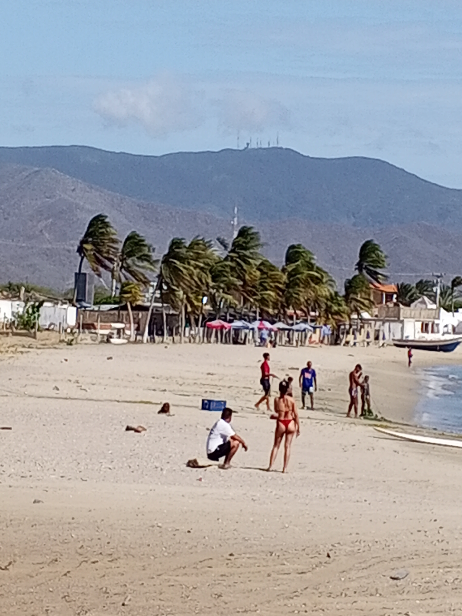 The image depicts a beach scene, likely in Juan Griego, Venezuela, given the location information. A mountain range is visible in the background, with some communication towers on the highest peak. Palm trees line the edge of the beach. In the foreground, a group of people is present. A man is squatting, while a woman in a bikini is standing nearby. Further away, other individuals are walking or interacting near the water's edge. A boat is beached near some buildings. Umbrellas and possible food stalls are seen lining the beach near the buildings. The sky is clear with light clouds.