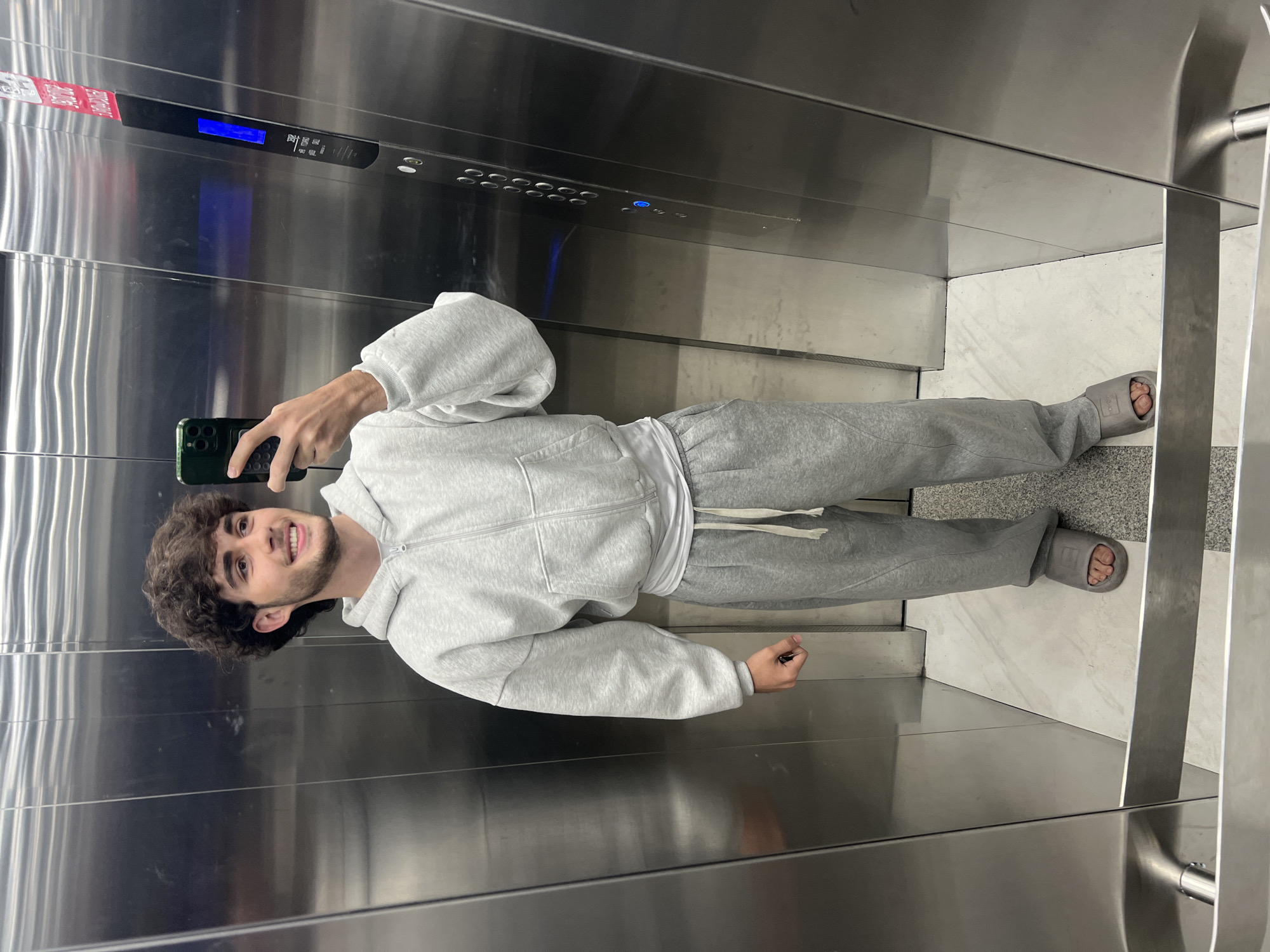 A young man takes a selfie in an elevator with stainless steel walls. He wears a gray hoodie and sweatpants, along with gray slide sandals. The elevator's control panel is visible above him. The floor is tiled in a light color. The location is likely in Yerevan, Armenia.