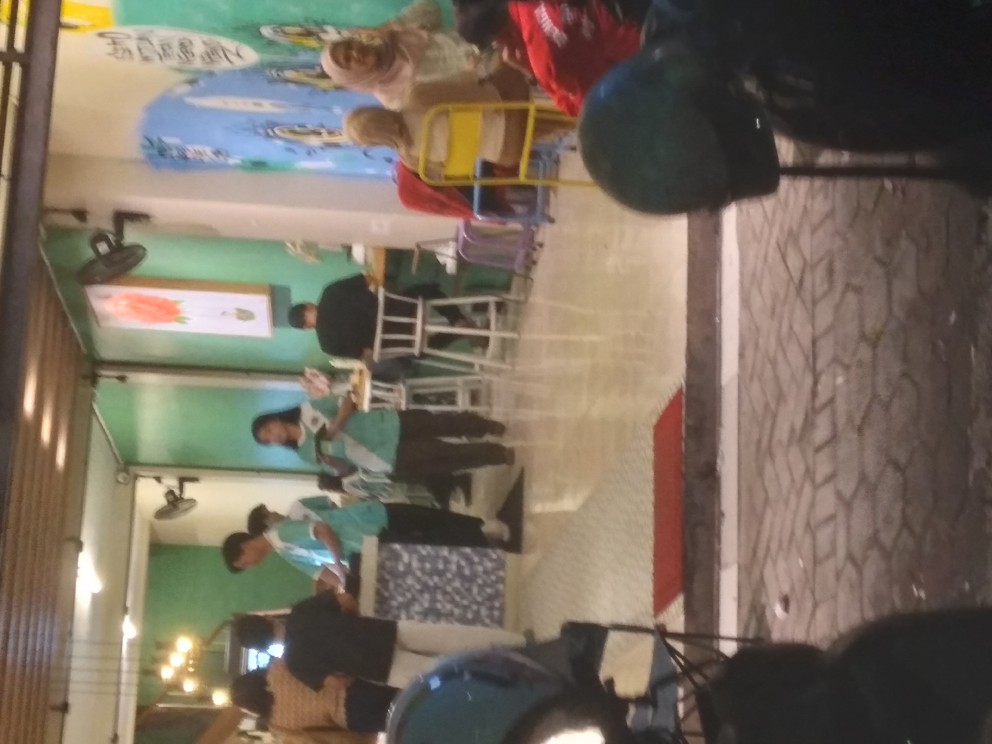 The image shows a group of people inside what appears to be a cafe or restaurant in Purwokerto, Indonesia. There are several individuals standing and sitting. In the foreground, a motorcycle helmet is visible. Inside the establishment, a woman in a green uniform is holding a tablet, possibly a server or cashier. Another person in a black shirt is seated at a table. Two individuals wearing light blue shirts are standing behind a counter area. In the background, a woman wearing a hijab is visible. The environment includes tables, chairs, and wall decorations, including a mural and a framed picture of a flower. The flooring is tiled. The lighting suggests it is evening.