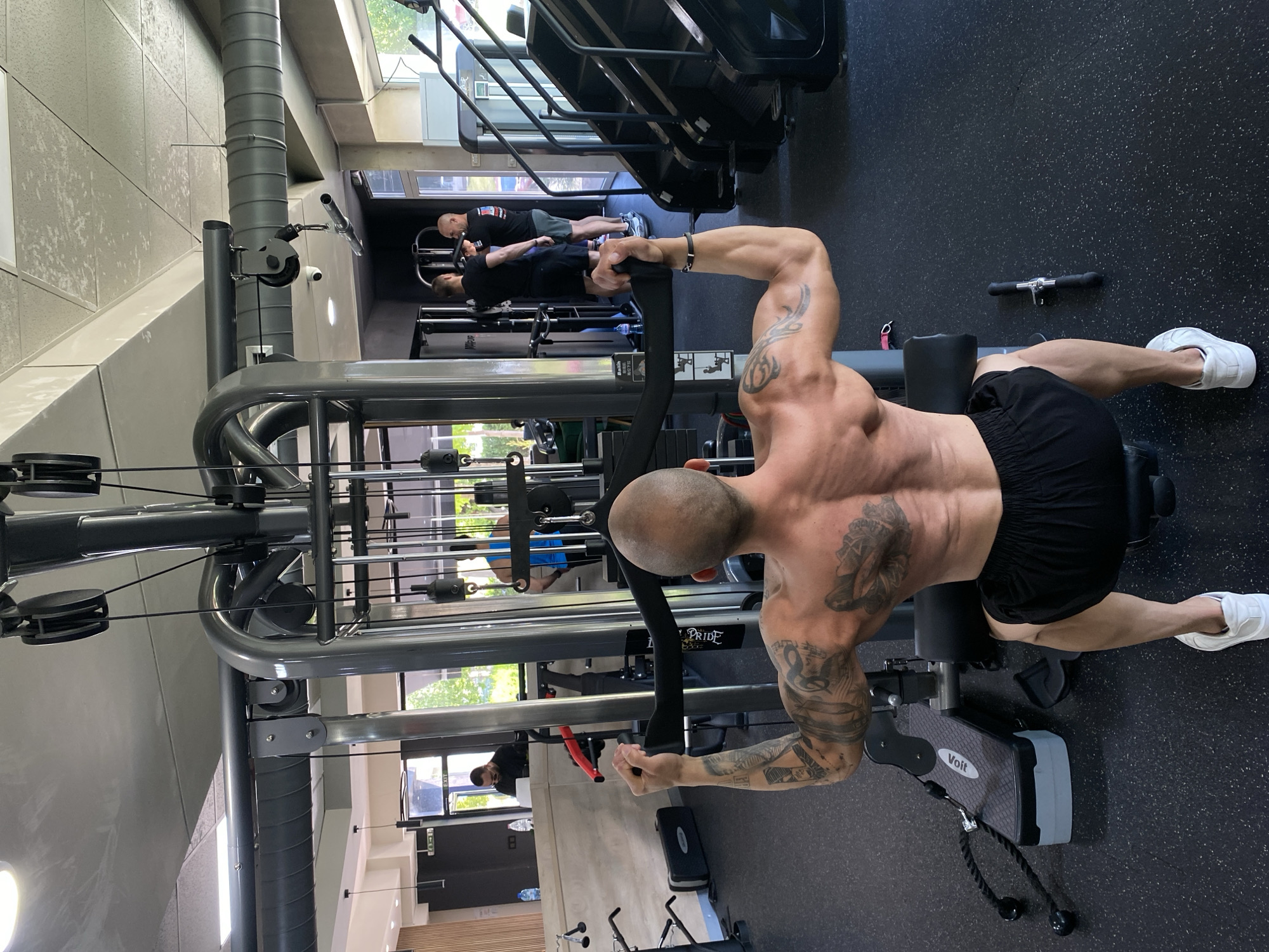 A man is working out on a seated cable row machine at a gym. The man is bald and has multiple tattoos on his arms and chest. He is wearing black shorts and white sneakers. In the background, two other people are working out on a treadmill and another piece of gym equipment. The gym appears to be located in Pleven, Bulgaria, based on visible details such as logos.