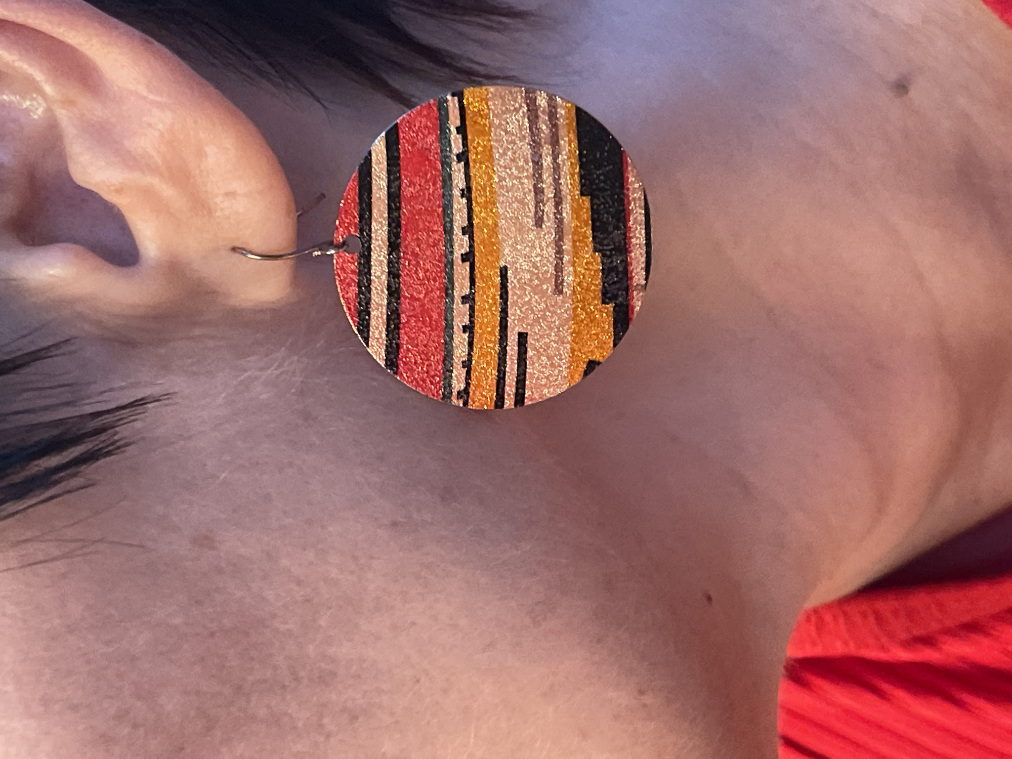 A close-up image shows a person's ear wearing a large, circular earring. The earring has a multicolored, striped pattern. The colors include red, orange, yellow, and black, with some lighter tan or beige stripes as well. The earlobe is pierced with a simple wire hook earring. The person has dark hair visible above the ear. Skin tone is light and fair. A red garment is visible at the bottom right corner of the frame. A mole is visible on the neck area of the person.