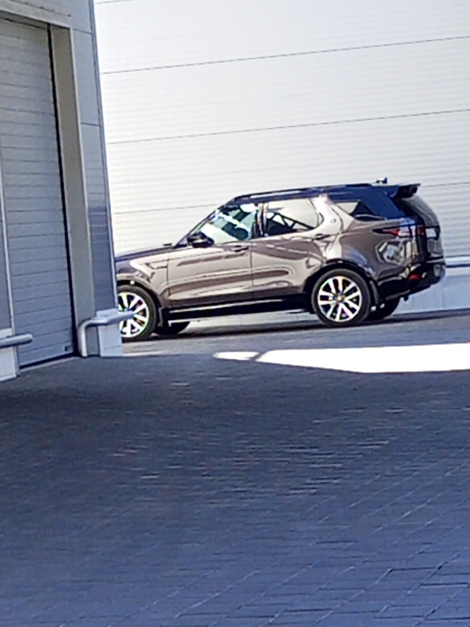 A dark-colored SUV is parked on a paved surface next to a large industrial-style garage door. The SUV is a Land Rover Discovery, as indicated by its distinctive rear design and roof spoiler. The vehicle is positioned at an angle, facing away from the camera. The garage door is made of corrugated metal and is mostly closed, with a portion of its upper section visible. The environment appears to be an outdoor urban setting, possibly a commercial or industrial area in Ceadîr-Lunga, Moldova. The ground is paved with dark gray interlocking bricks. The lighting suggests it is daytime, with sunlight casting shadows. There are no people visible in the image.