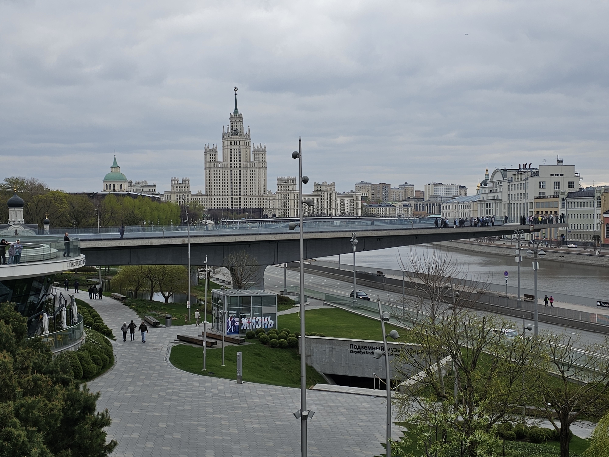 The image depicts a cityscape in Moscow, Russia, under an overcast sky. The dominant feature is the Kotelnicheskaya Embankment Building, one of the Seven Sisters skyscrapers, centrally positioned in the background. A modern pedestrian bridge spans the scene horizontally, crowded with people. Below the bridge flows the Moscow River.
In the foreground, a paved area leads towards a park entrance identified as "Zaryadye Underground Museum." People are walking along the paths. Trees are in various stages of budding. Streetlights line the walkways and roads. Cars are visible on the road parallel to the river. The image captures an urban scene with a mix of modern infrastructure and historical architecture.