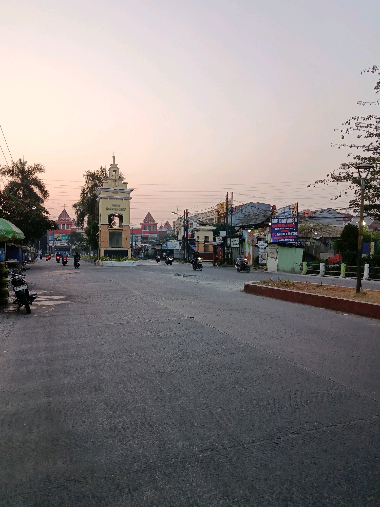 The image depicts a street scene in Bekasi, Indonesia, likely during dawn or dusk, given the soft, diffused light. A prominent landmark in the center is a tall, square tower with architectural detailing and a cross on top, identified by signage as "Taman Harapan Baru". This landmark is situated at an intersection.
The street is paved with asphalt and appears to be moderately busy with several motorcycles in motion. On the left side of the road, a green and white umbrella shades a small stall, and a motorcycle is parked. Palm trees and other vegetation are visible, along with utility poles and overhead wires.
Buildings line the sides of the street, exhibiting a mix of commercial and residential architecture. Several signs are visible, including one for "SKP CARWASH" and "ABILITY MOTOR". The overall environment suggests a typical urban or suburban setting in Indonesia. The sky is a pale orange and pink, indicating the time of day.