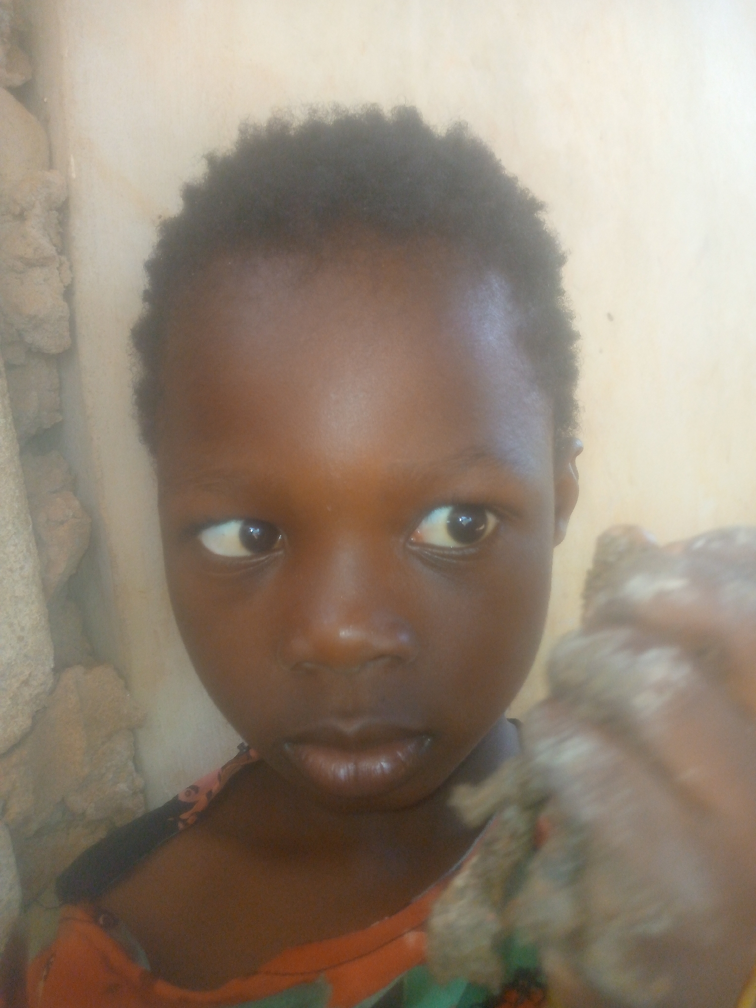 A close-up image shows a young, dark-skinned child with short, dark hair. The child is looking slightly to the left of the frame. The background consists of a beige wall on the right and a gray, textured surface (possibly a stone or concrete wall) on the left. In the lower right of the frame is an out-of-focus object held in the child's hand. The lower portion of the child's body is partially visible, with what appears to be an orange and black pattern on the child's shirt. The location is not identifiable as Unknown, Unknown from the image content.