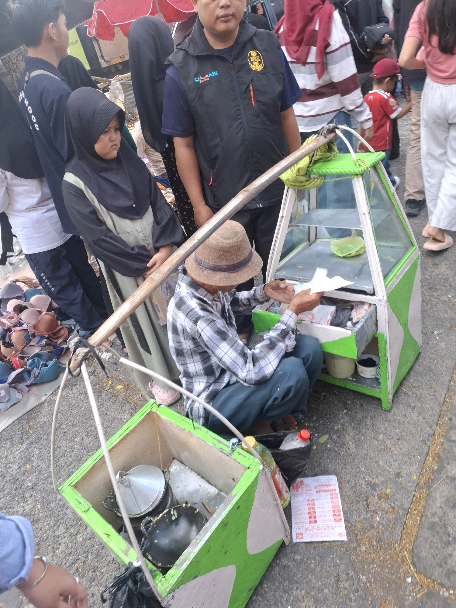 This image captures a street scene in Karangtengah, Indonesia, likely a marketplace or busy thoroughfare. In the foreground, a man wearing a plaid shirt and a hat crouches beside a small, triangular food cart. The cart, painted with green and white camouflage patterns, has a glass display case on top and a drawer below filled with various items. The man is holding a piece of paper.
To the left of the food cart, a young child in a black hijab sits with her head down. Behind her, a person wearing a blue uniform with white lettering and a black vest is visible. Further back, other individuals, including a child in a red shirt and cap, are present. The ground is paved with asphalt and scattered with debris. A pile of brassieres is visible on the ground to the far left. The overall impression is of a bustling commercial area with various goods and people interacting.