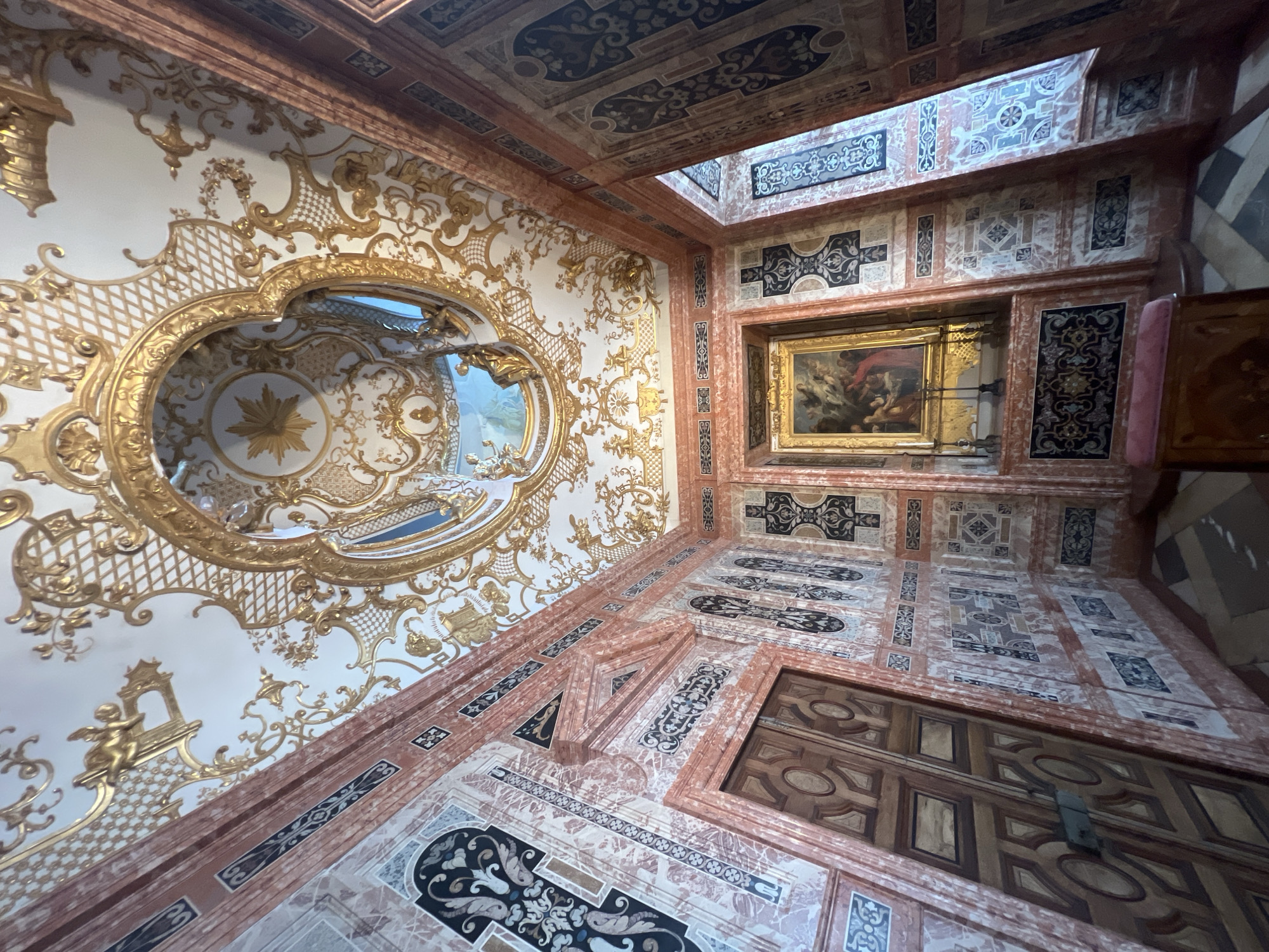 This image depicts an ornate room, likely in a palace, featuring a highly decorated ceiling and walls. The ceiling is white with elaborate gold stucco work, including cherubic figures, scrolling patterns, and a central oval inset containing a painted scene. The walls are a combination of pink marble-like material and intricate dark patterns, with framed panels containing geometric and floral motifs. A wooden door with an oval inset is visible on the left, and a framed painting depicting a religious or mythological scene hangs on the right wall, above a small altar with candlesticks. A padded stool or bench is in the foreground. The floor is tiled in a checkered pattern of light and dark squares. The scene suggests a lavish interior decoration, characteristic of Baroque or Rococo architecture. This room is located in the New Palace (Neues Schloss) in Oberschleißheim, Germany, part of the Schleissheim Palace complex. There are no people or animals present in the image.