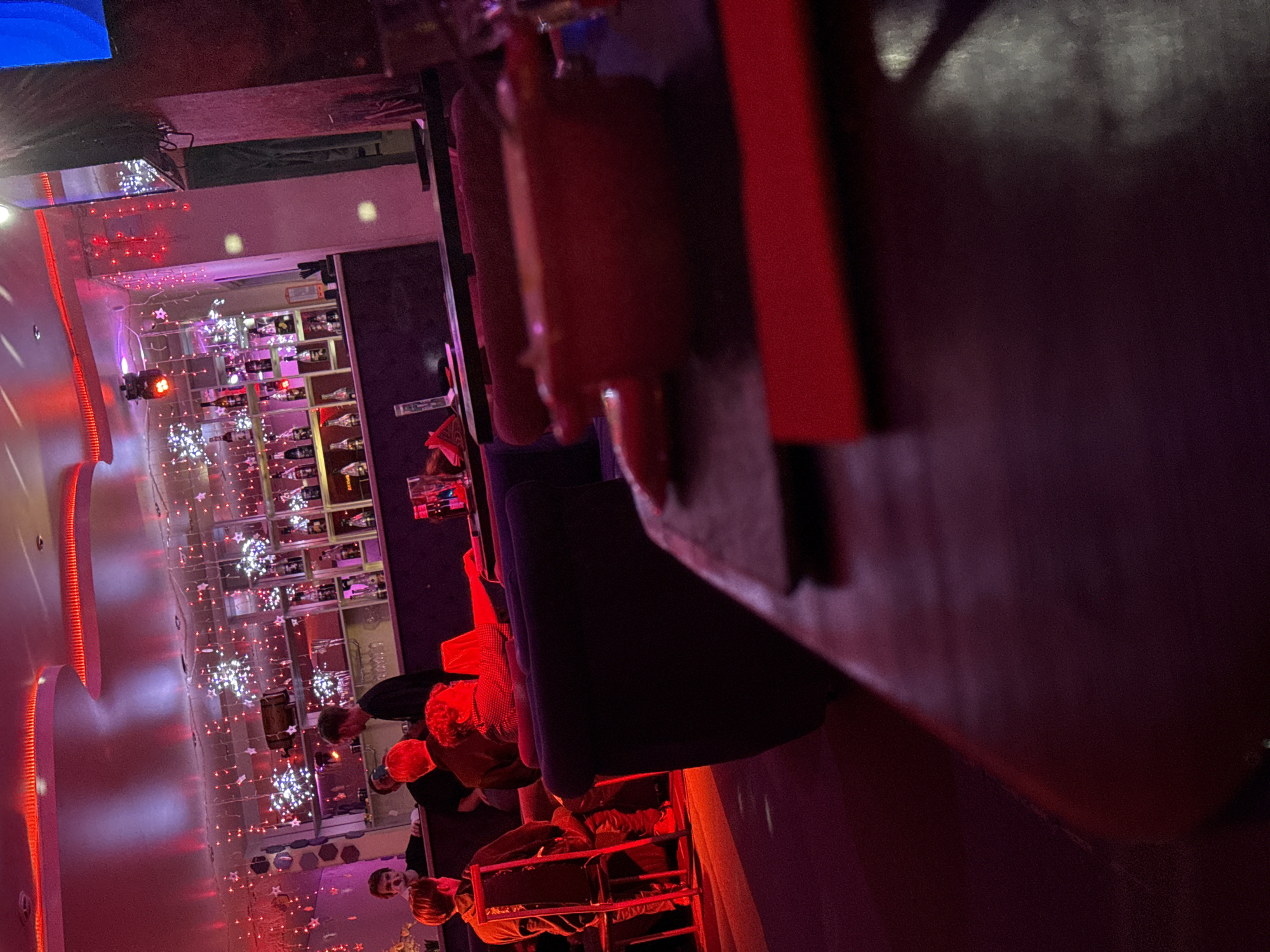 The image captures a dimly lit interior space, likely a bar or nightclub, with a red and purple lighting scheme. A bar shelf displays numerous bottles of alcohol. Several people are seated at tables and at the bar. Christmas-themed decorations, including string lights and star ornaments, are visible in the background. The location could be Votkinsk, Russia, given the prompt, but no distinct local landmarks are visible in the image. The photograph is taken from an obstructed perspective, with part of the bar or table dominating the right foreground.